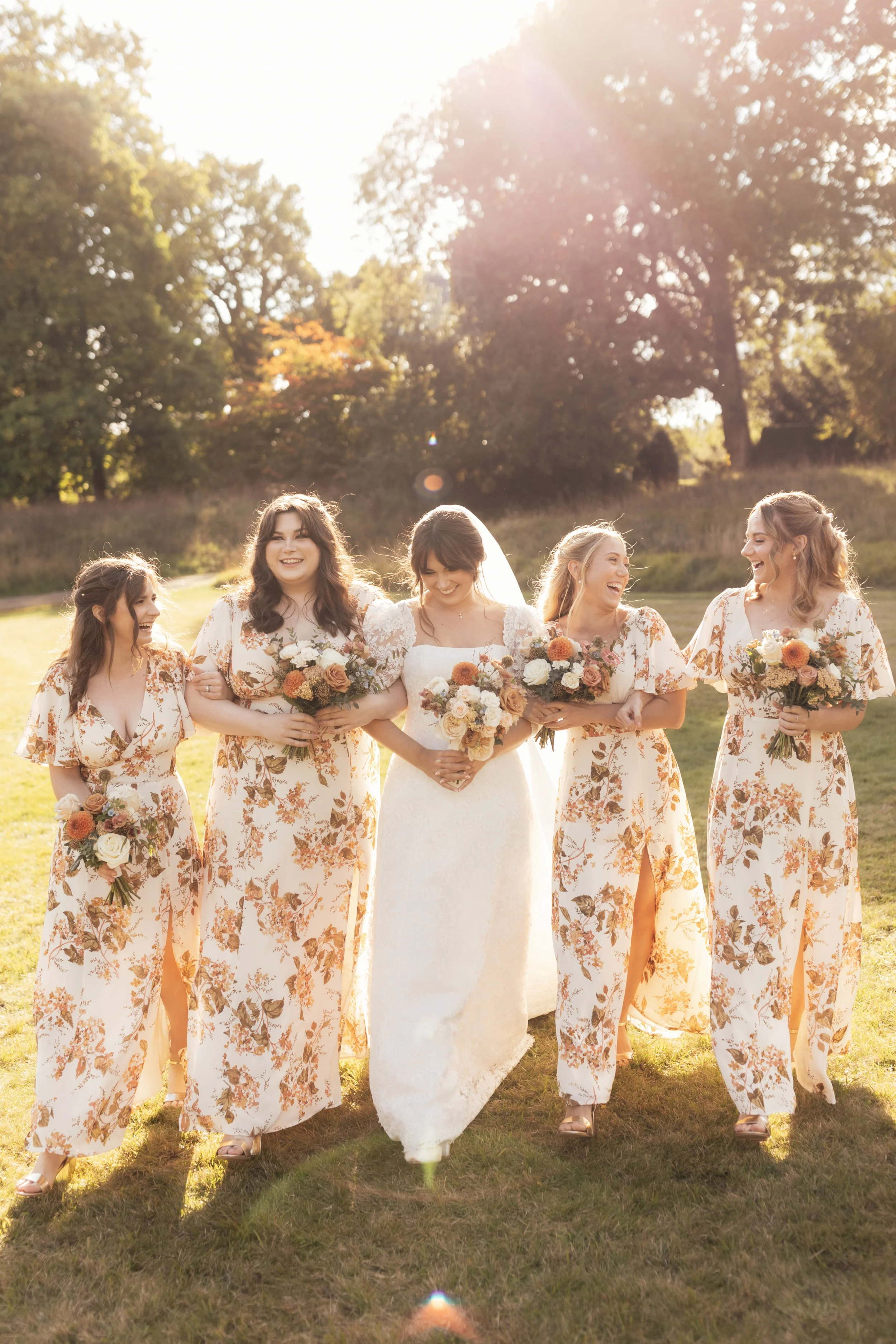 Group of five women in wedding dresses and matching floral robes holding bouquets, walking outdoors on grass field with trees and sunlight in the background, during sunset.