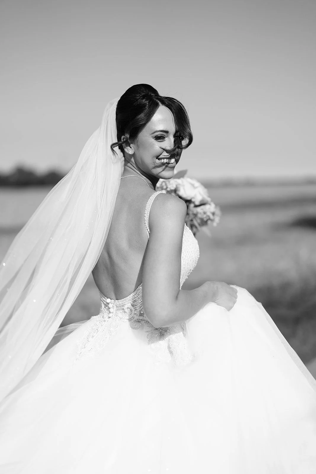 Black and white photo of a smiling bride in a wedding dress, holding a bouquet, outdoors on a sunny day.