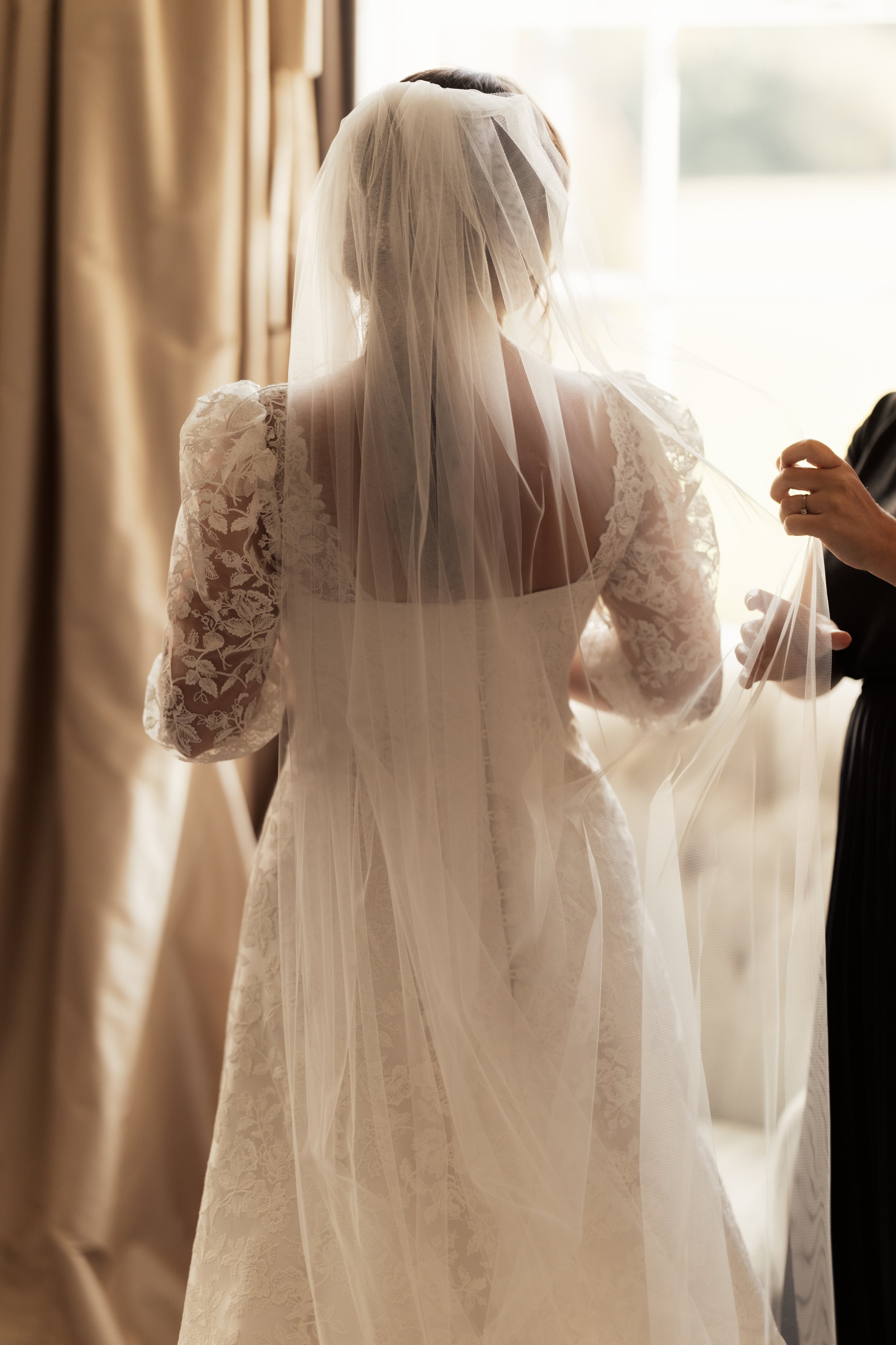 A bride wearing a white lace wedding dress and veil, standing near a window with sunlight streaming in, beside beige curtains.