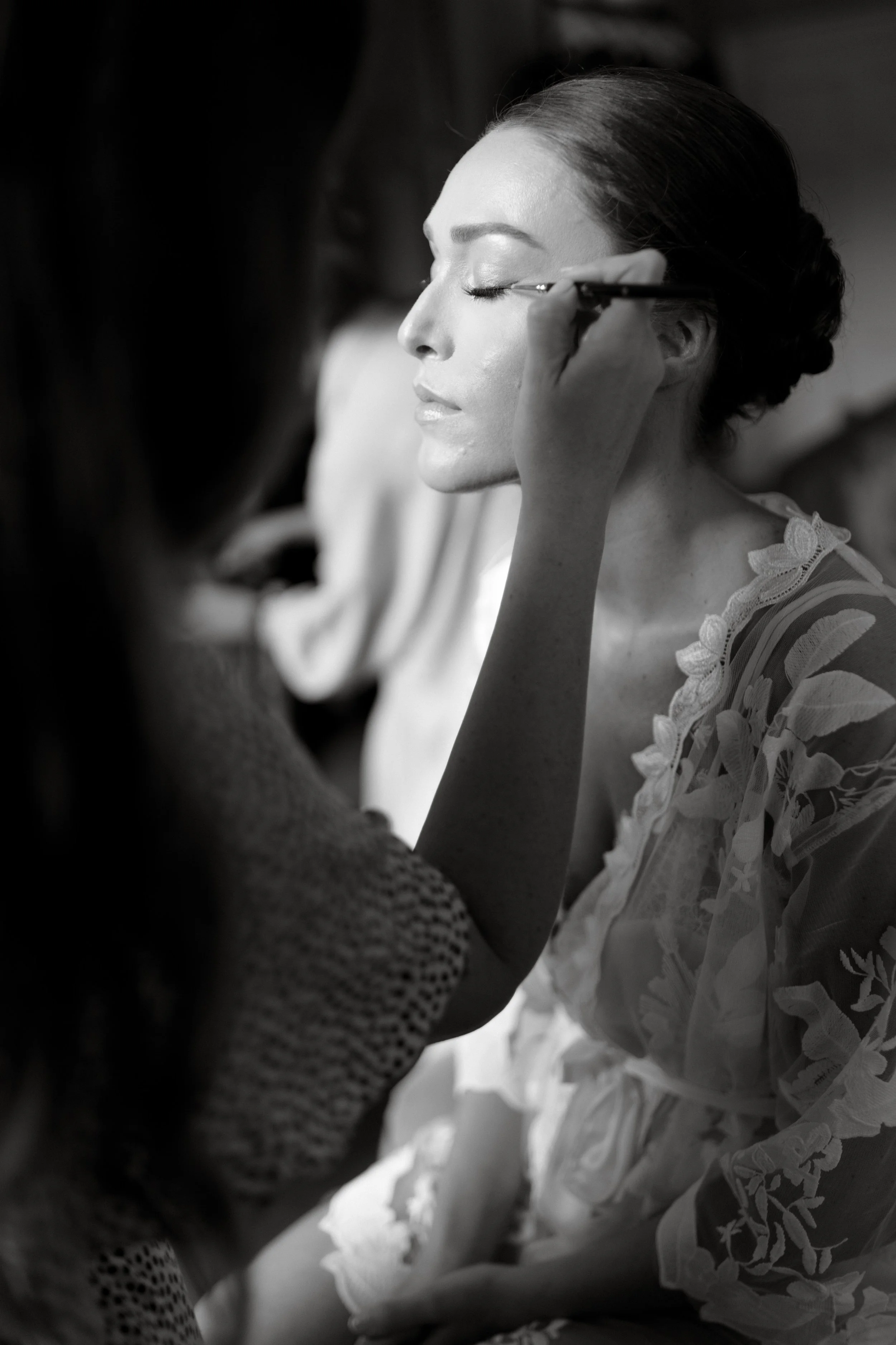 A woman with closed eyes getting makeup applied with a brush by a makeup artist. She is wearing a lace dress with floral embroidery.