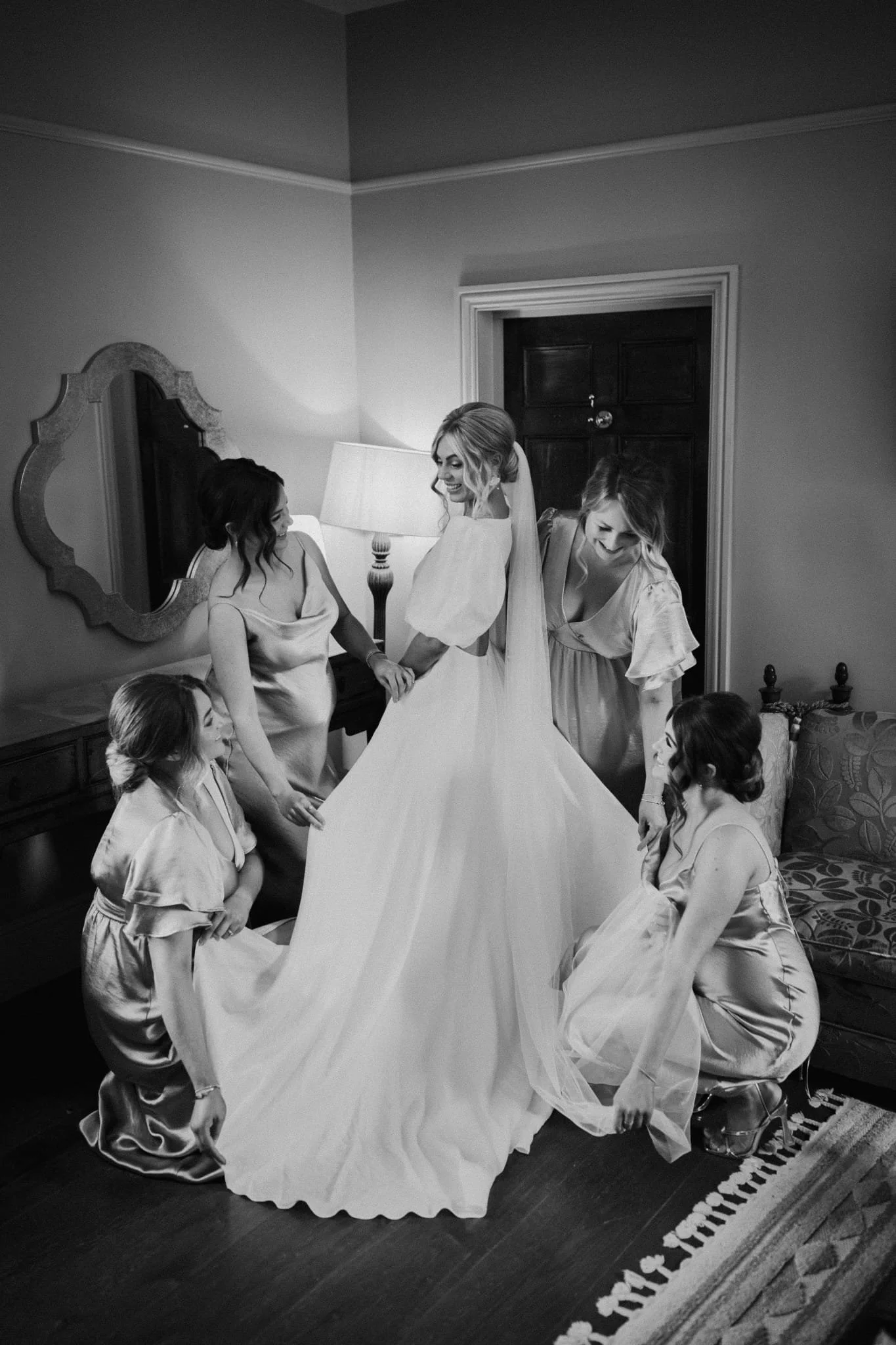 A bride in a wedding gown holding hands with a bridesmaid in a robe, surrounded by five girls in matching robes, all kneeling or sitting around her on a rug in a bedroom.