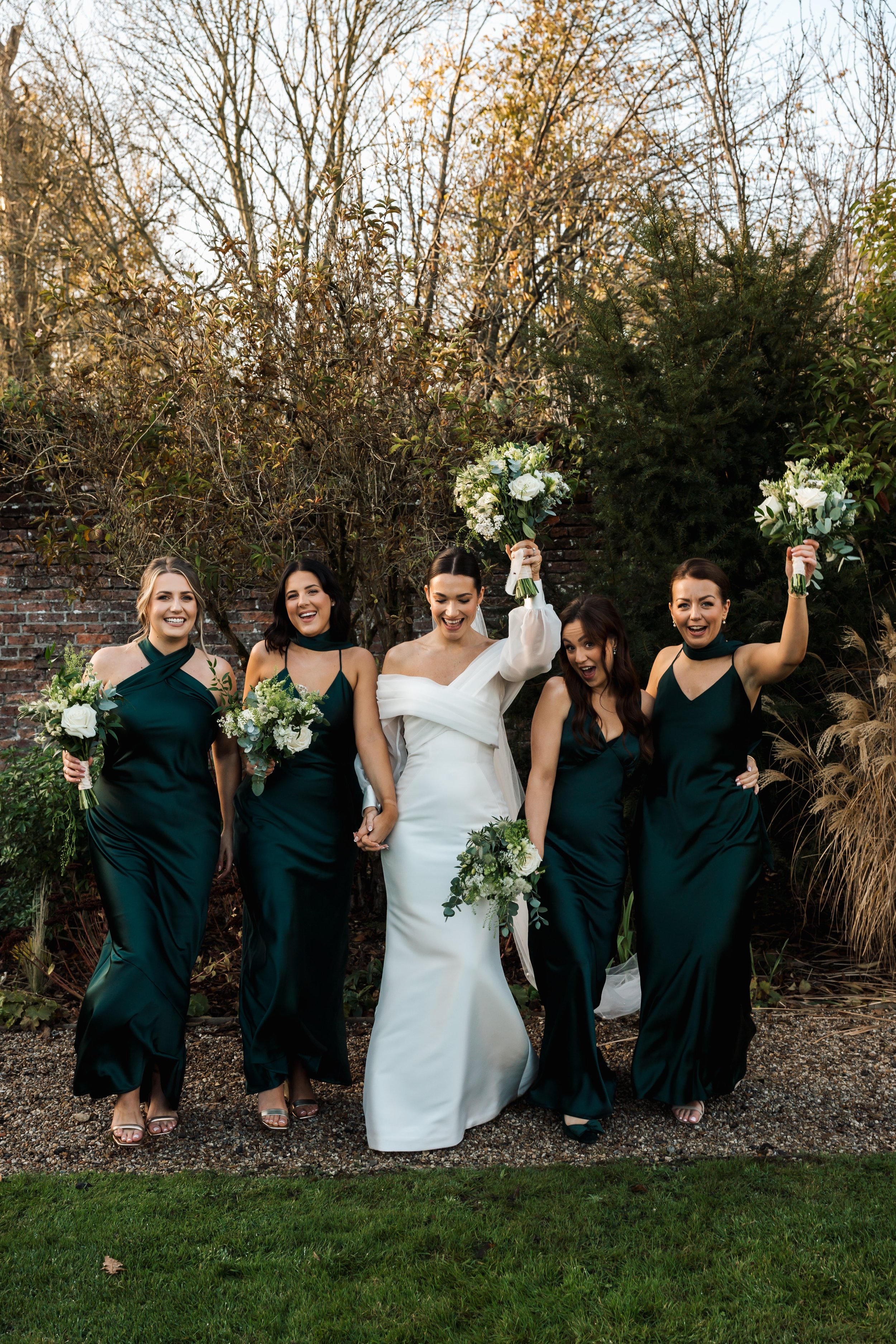 A bride and four bridesmaids celebrating outdoors, holding bouquets and raising their flowers, with trees and a brick wall in the background.