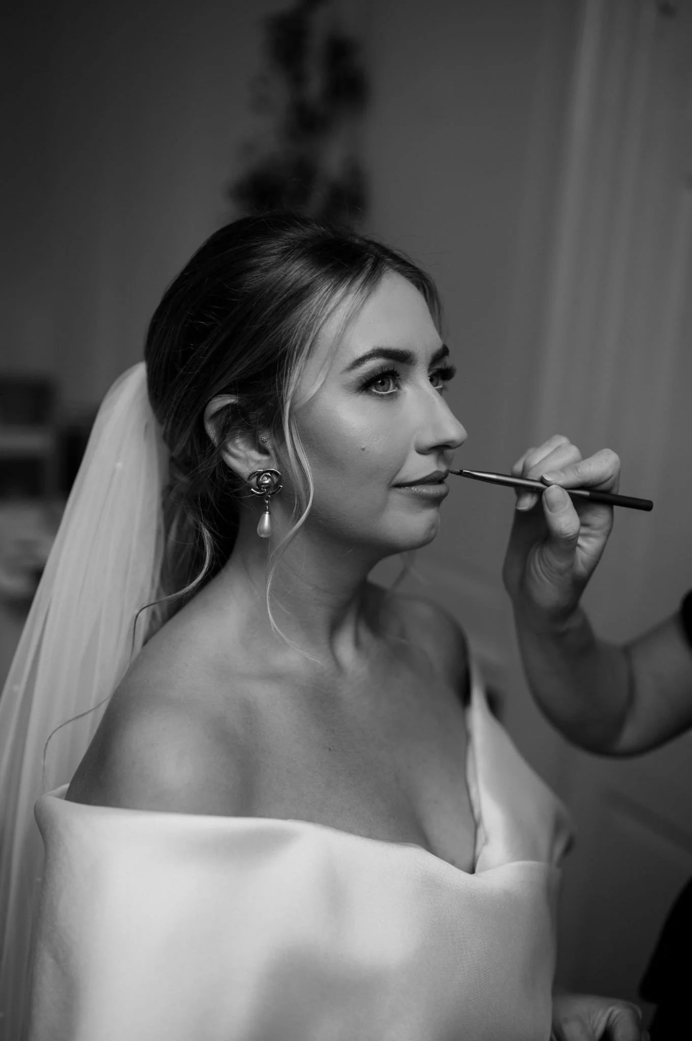 A bride with styled hair and earrings is having makeup applied to her lips by a makeup artist, with a veil draped over her shoulder, in a black and white photo.