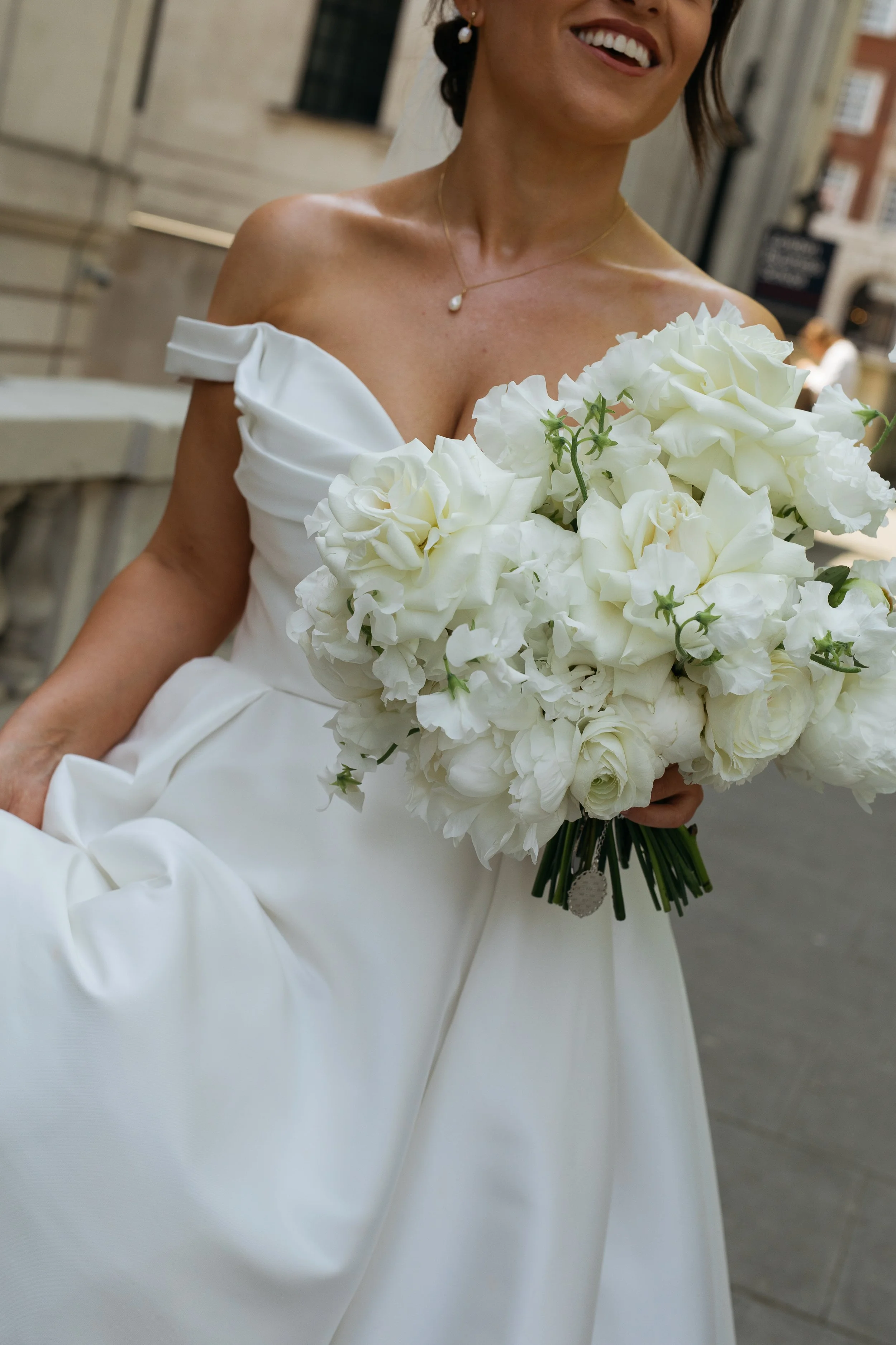 A woman in a white wedding dress holding a large bouquet of white flowers, smiling with her teeth showing.