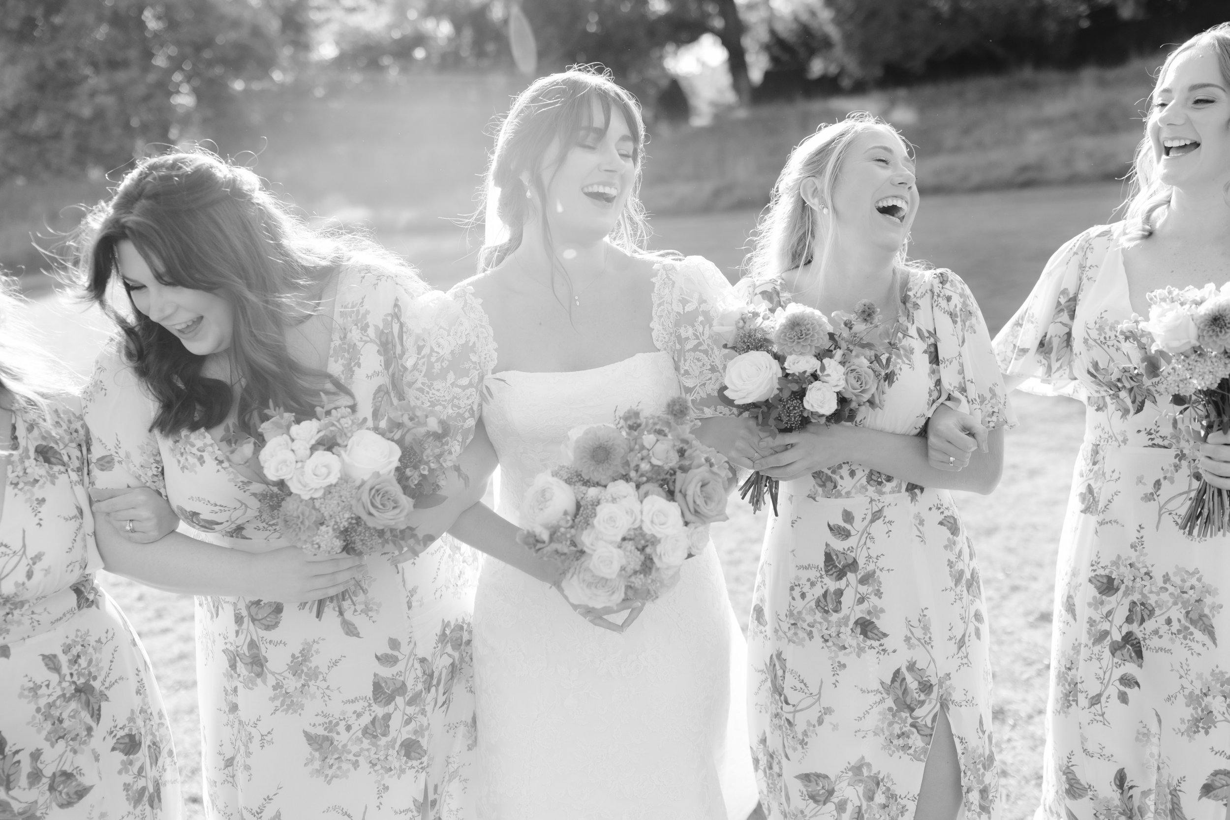 Black and white photo of a bride and bridesmaids holding flowers and laughing outdoors.