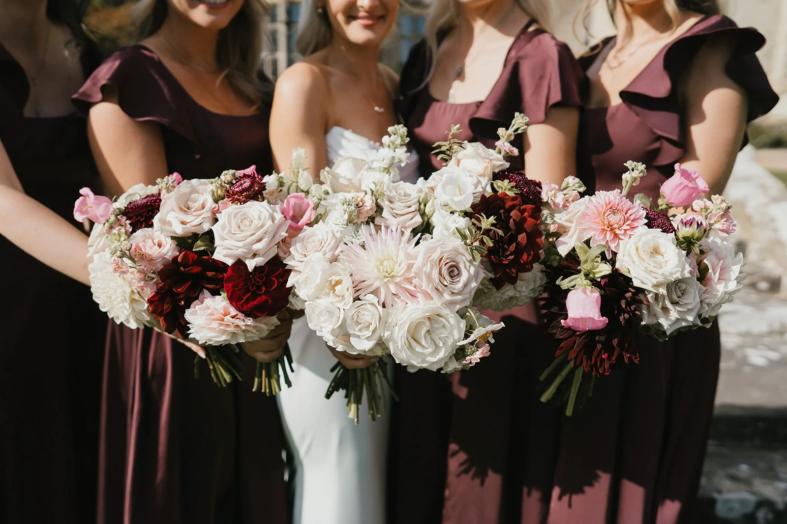 Group of women holding large bouquets of mixed pink, white, and burgundy flowers, dressed in formal attire, at an outdoor wedding or event.