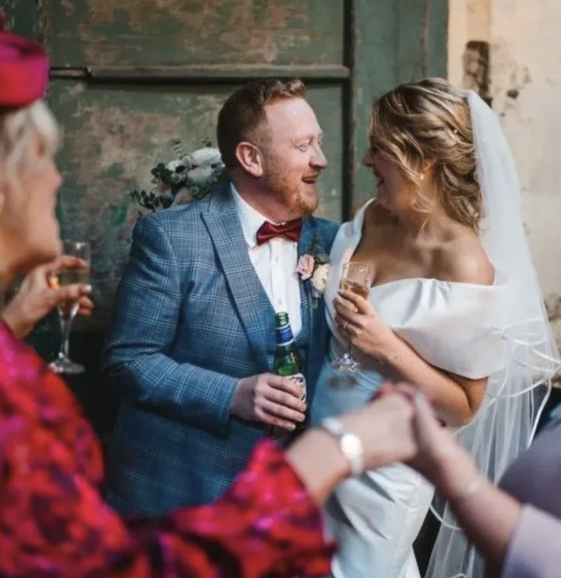 A bride and groom smiling at each other during a wedding celebration, holding drinks, with guests in the foreground.