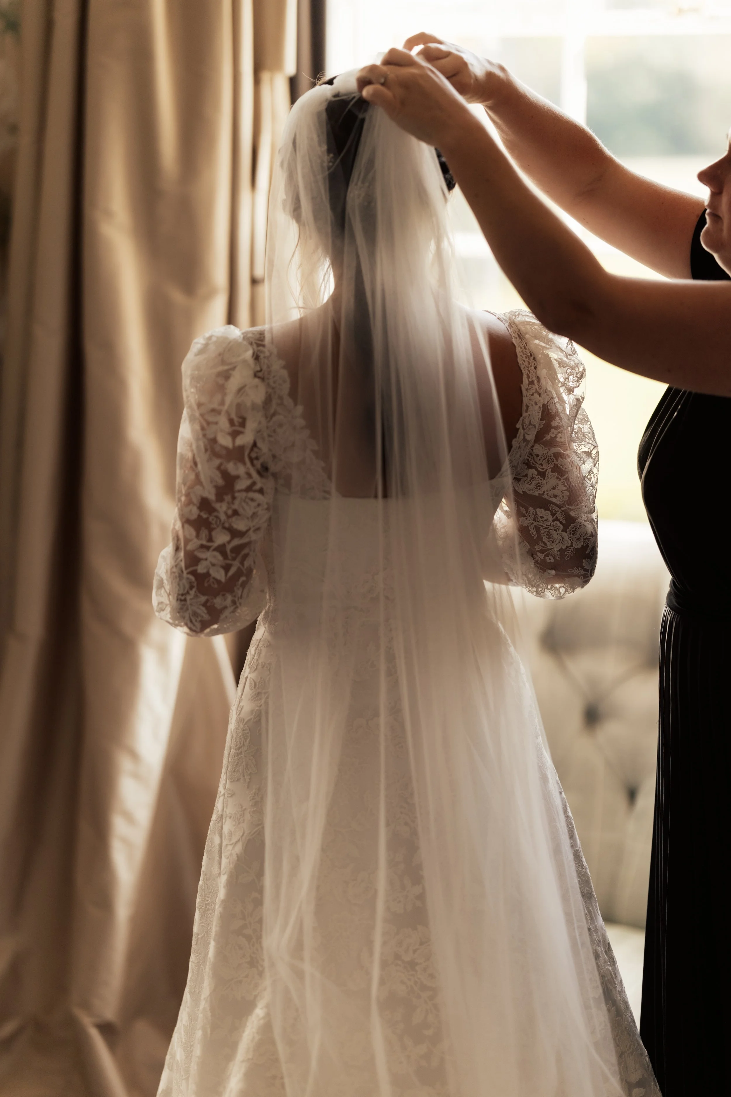A person helping a bride adjust her veil in a room filled with natural light.