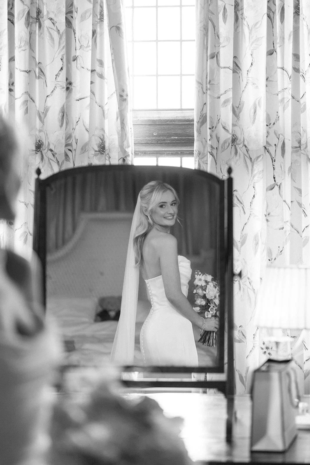 A black and white photo shows a bride in a wedding dress holding a bouquet, smiling as she looks into a mirror in a well-lit room with floral curtains and a window in the background.