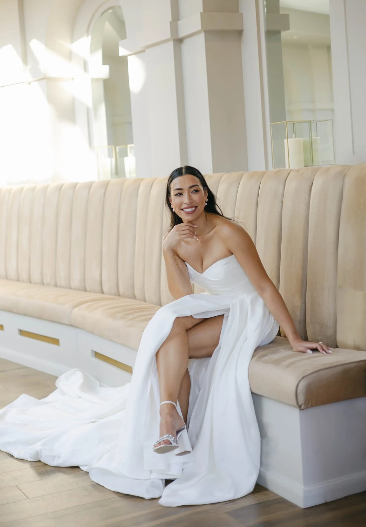 A woman in a white strapless dress sitting on a beige cushioned bench in a bright, elegant room. She is smiling, with one hand on her chin and her legs crossed. The room has tall windows, white walls, and candle decor.