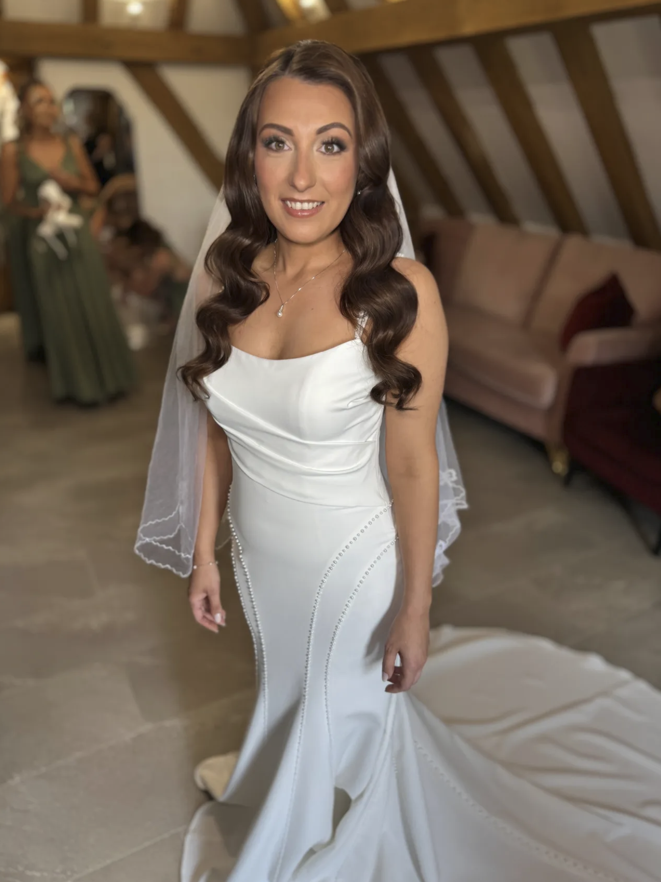 A smiling woman in a wedding dress with long, wavy brown hair standing in a room with wooden beams on the ceiling. In the background, there are two women, one in a green dress, and a sofa with pillows.