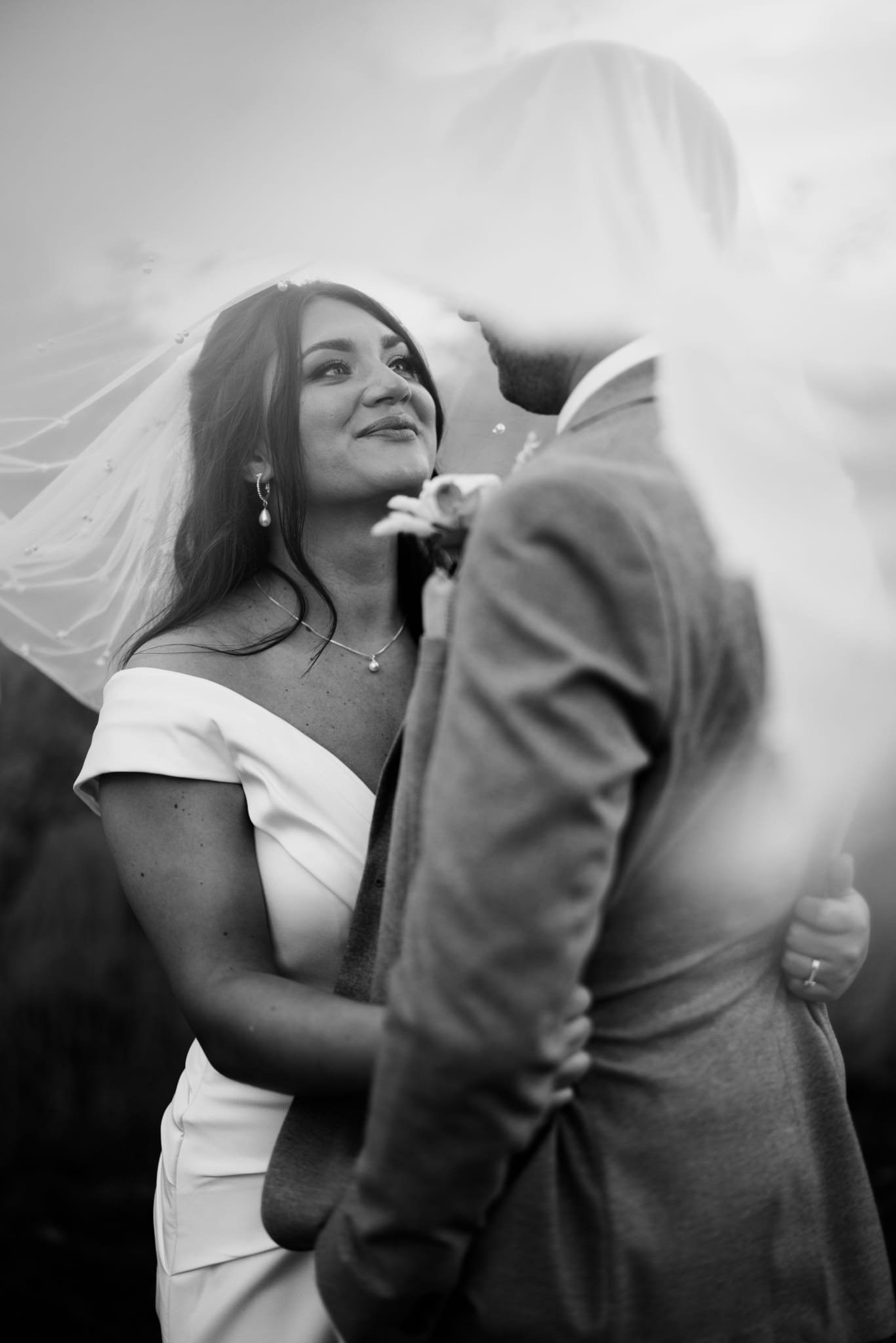 A black-and-white photo of a bride and groom on their wedding day, gazing into each other's eyes. The bride is wearing a white dress, pearl earrings, and a necklace, with a veil on her hair. The bride's hand is resting on the groom's chest, and he is in a suit. They are holding each other close, under a veil or fabric canopy.