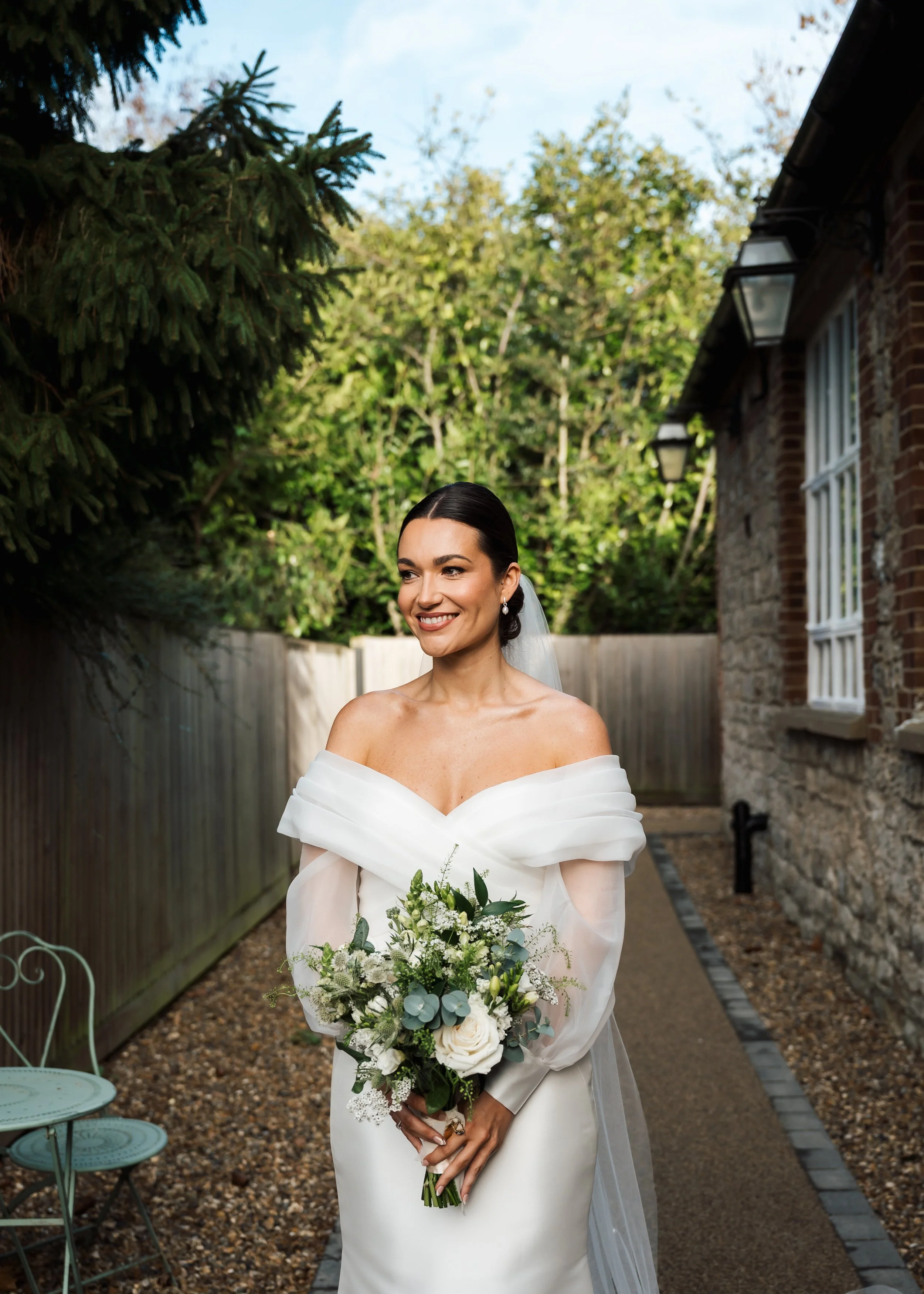 A bride in an off-shoulder white wedding dress with dark hair in an updo, holding a bouquet of white roses, greenery, and eucalyptus, standing outdoors near a brick house and lush trees.