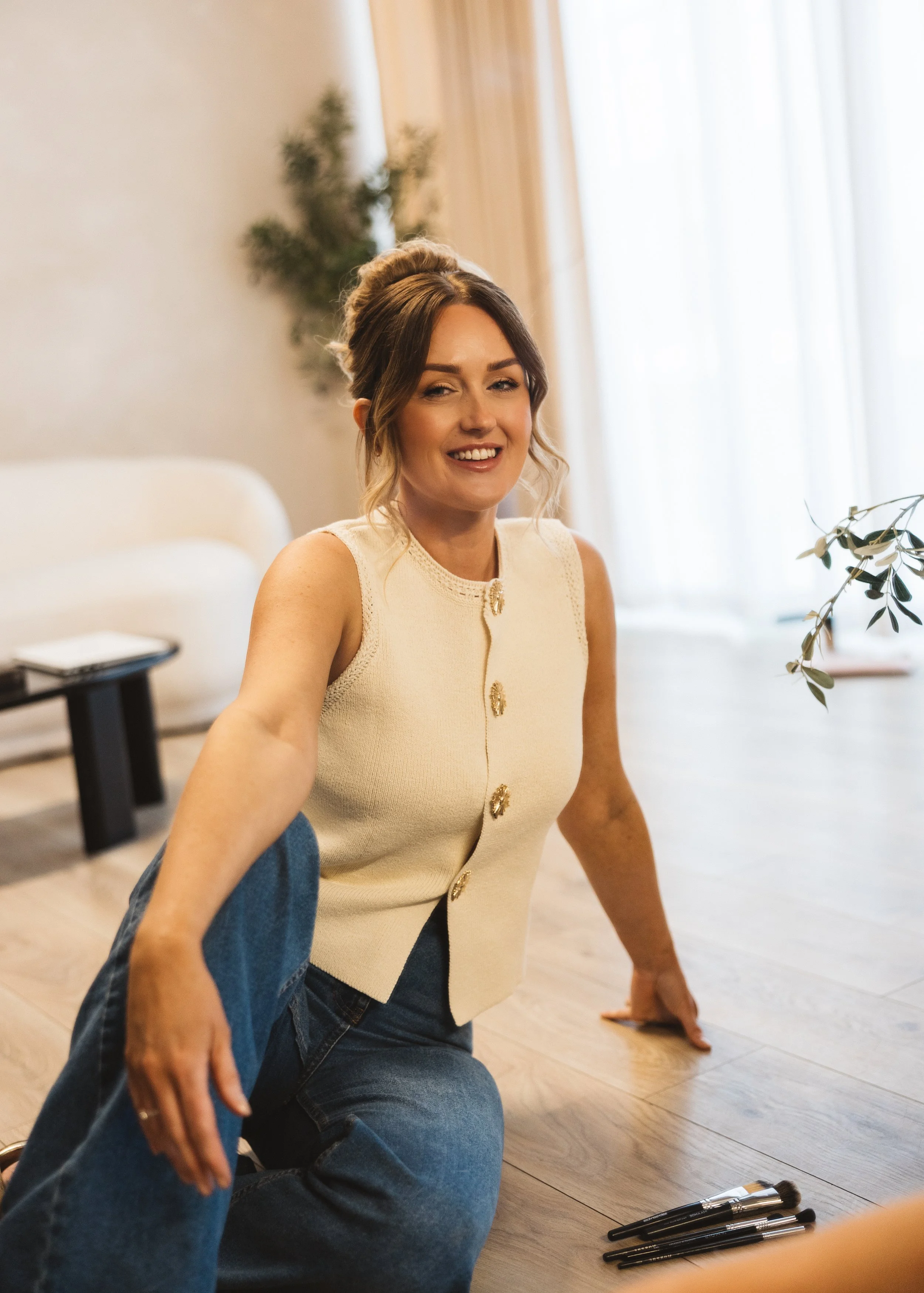 A woman smiling with a makeup brush in front of her, sitting on the floor in a bright room with a window, a sofa, a side table, and makeup brushes on the floor.