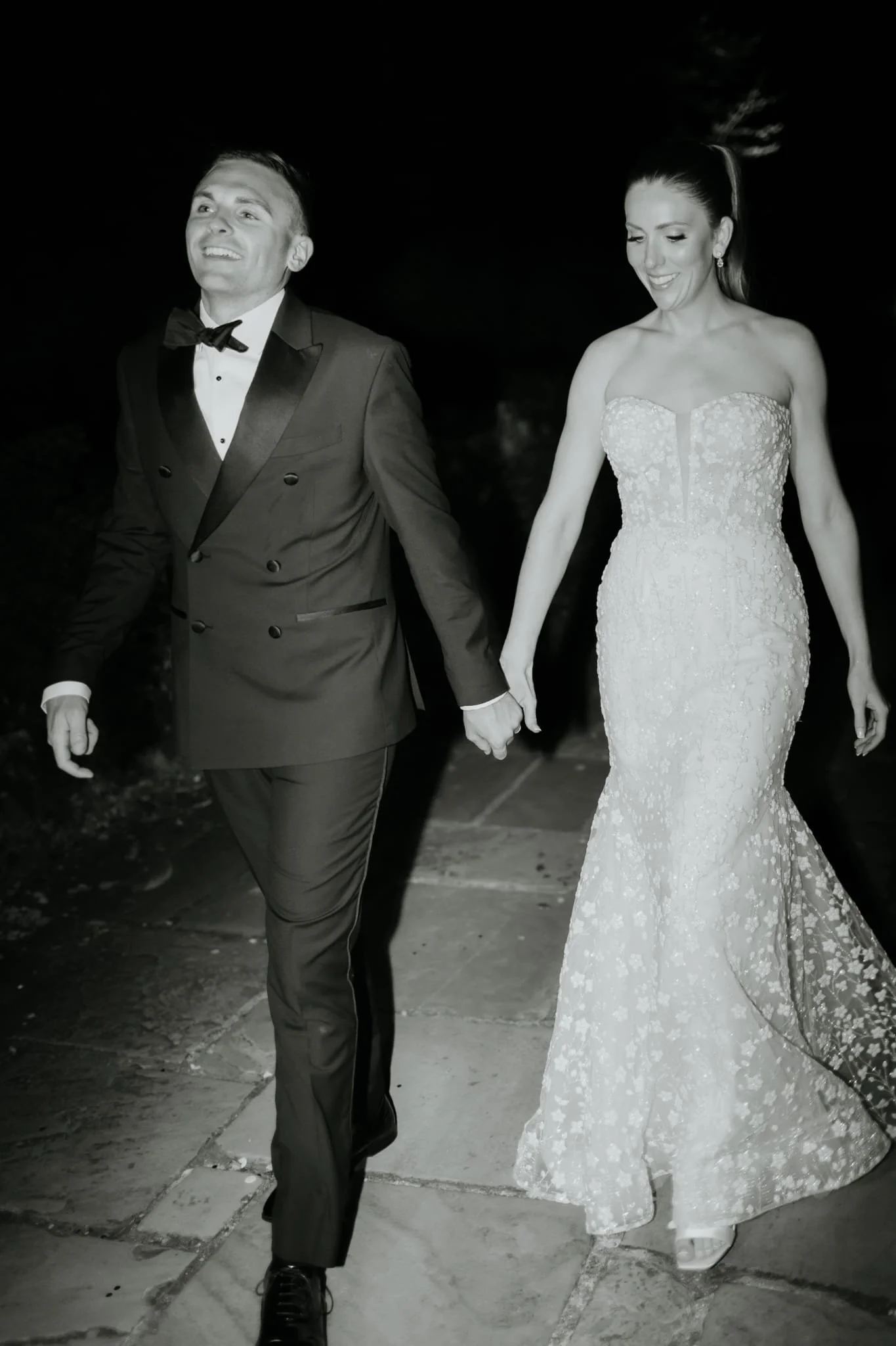 A black and white photo of a man and woman holding hands, dressed in formal wedding attire. The man is wearing a tuxedo with a bow tie, and the woman is in a strapless wedding gown with lace details. They are walking on a sidewalk, smiling and lookin