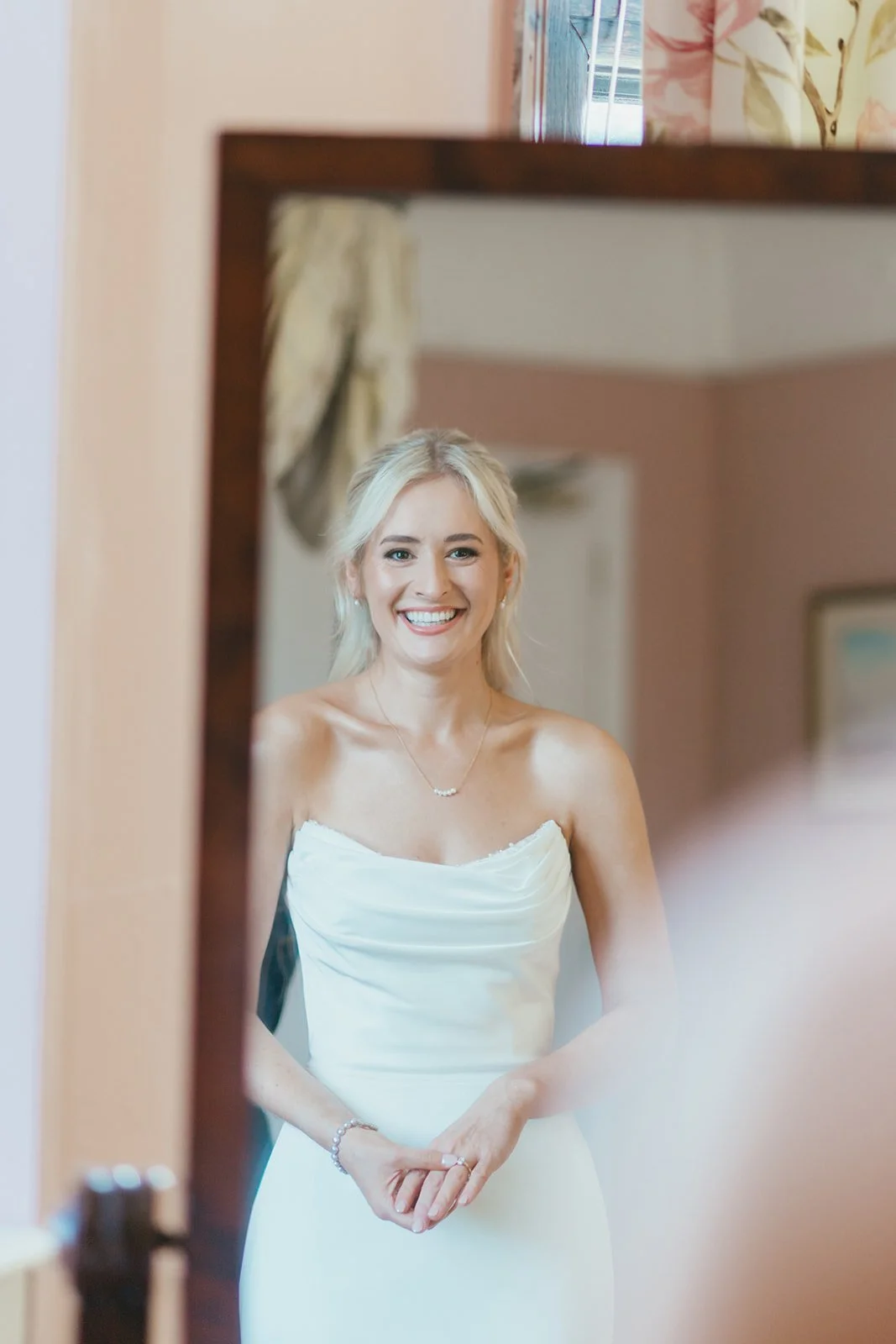 A smiling woman in a white dress taking a selfie in a mirror.
