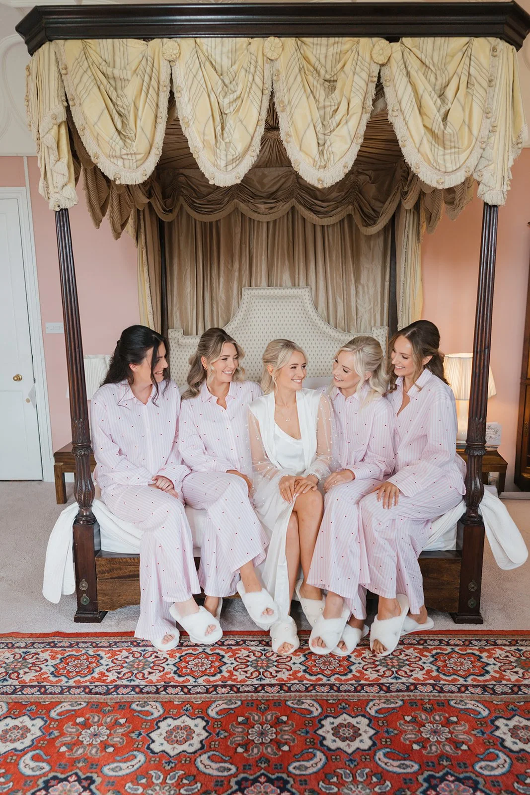Group of five women sitting on a bed in pajamas, smiling and looking at each other, in a bedroom with a canopy, pink walls, and a patterned rug.