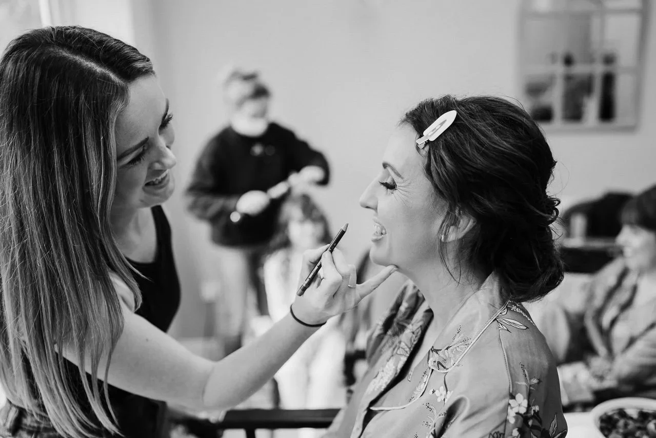 A woman is applying makeup to another woman with a makeup brush in an indoor setting, with two other women in the background.