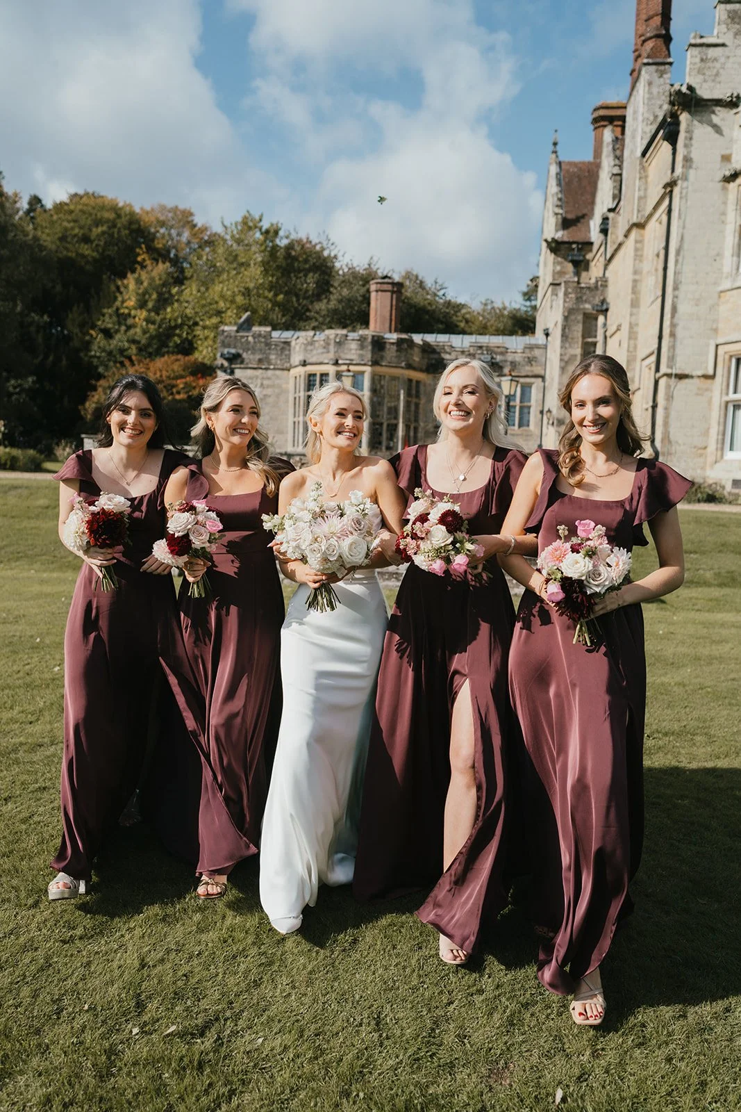 Bride and five bridesmaids walking on a lawn in front of a historic stone building on a sunny day, all holding bouquets of flowers.