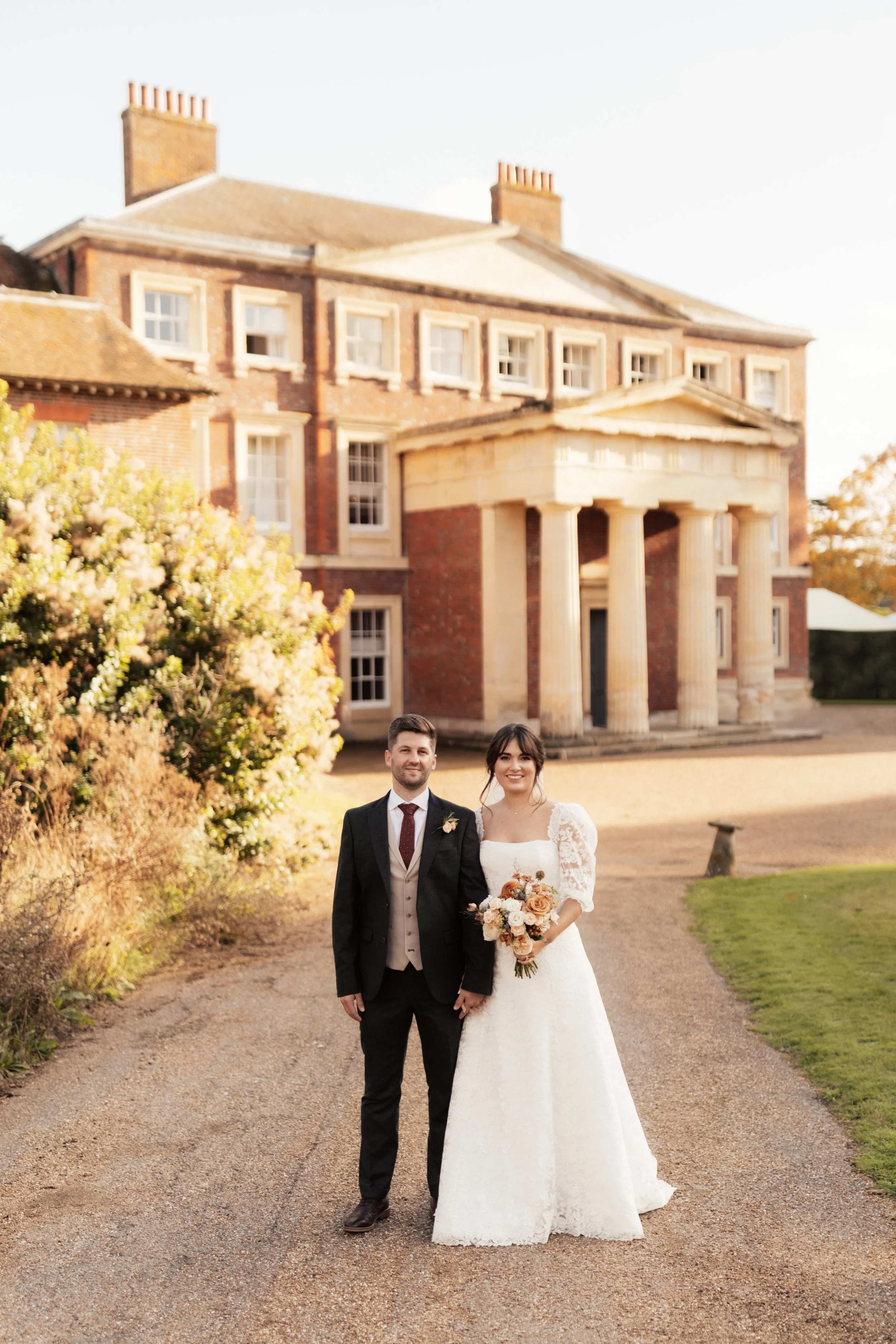 A newlywed couple stands on a gravel path outside a large, historic brick house with classical columns. The groom is in a black suit with a vest and tie, and the bride is in a white lace wedding gown holding a bouquet of flowers. The house has multip