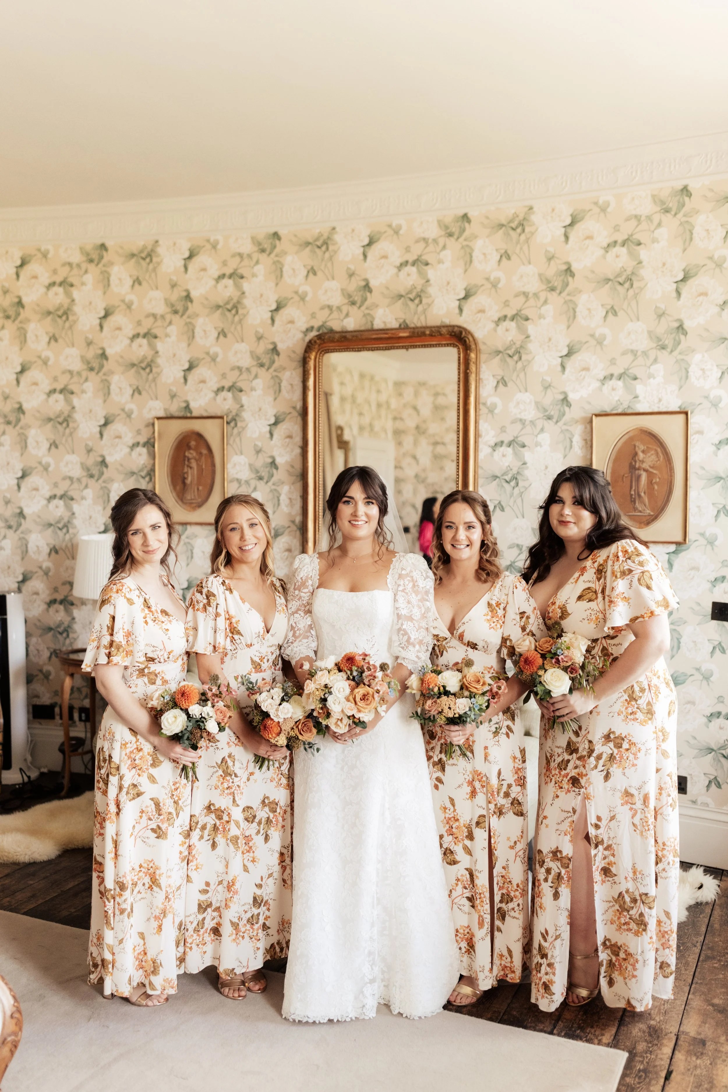 Bride and five bridesmaids, dressed in matching floral pajamas, standing together in a decorated room, holding bouquets, with a mirror and floral wallpaper in the background.