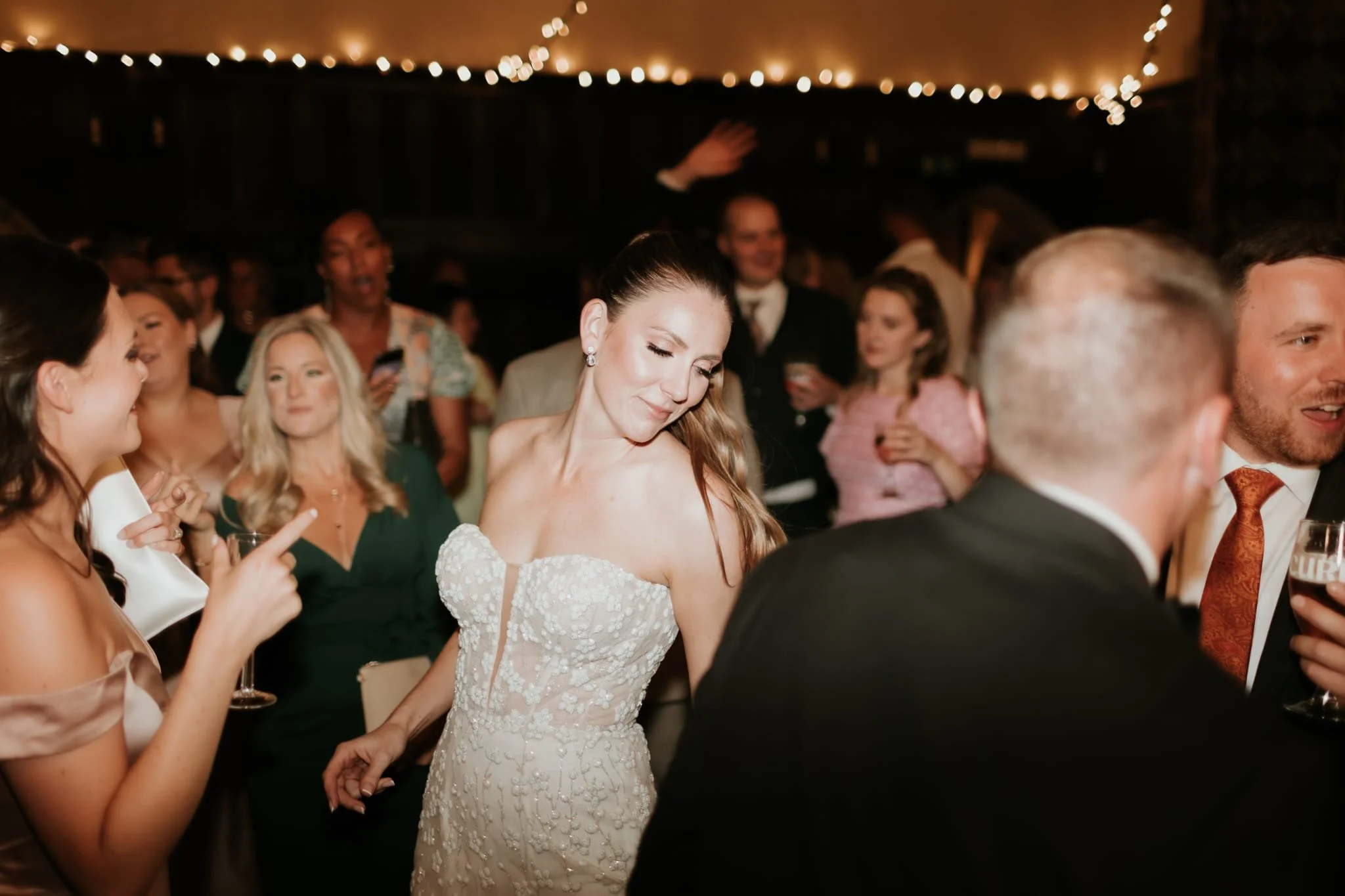 A bride in a white wedding dress with lace details dancing at a wedding reception surrounded by guests in formal attire, with string lights overhead.
