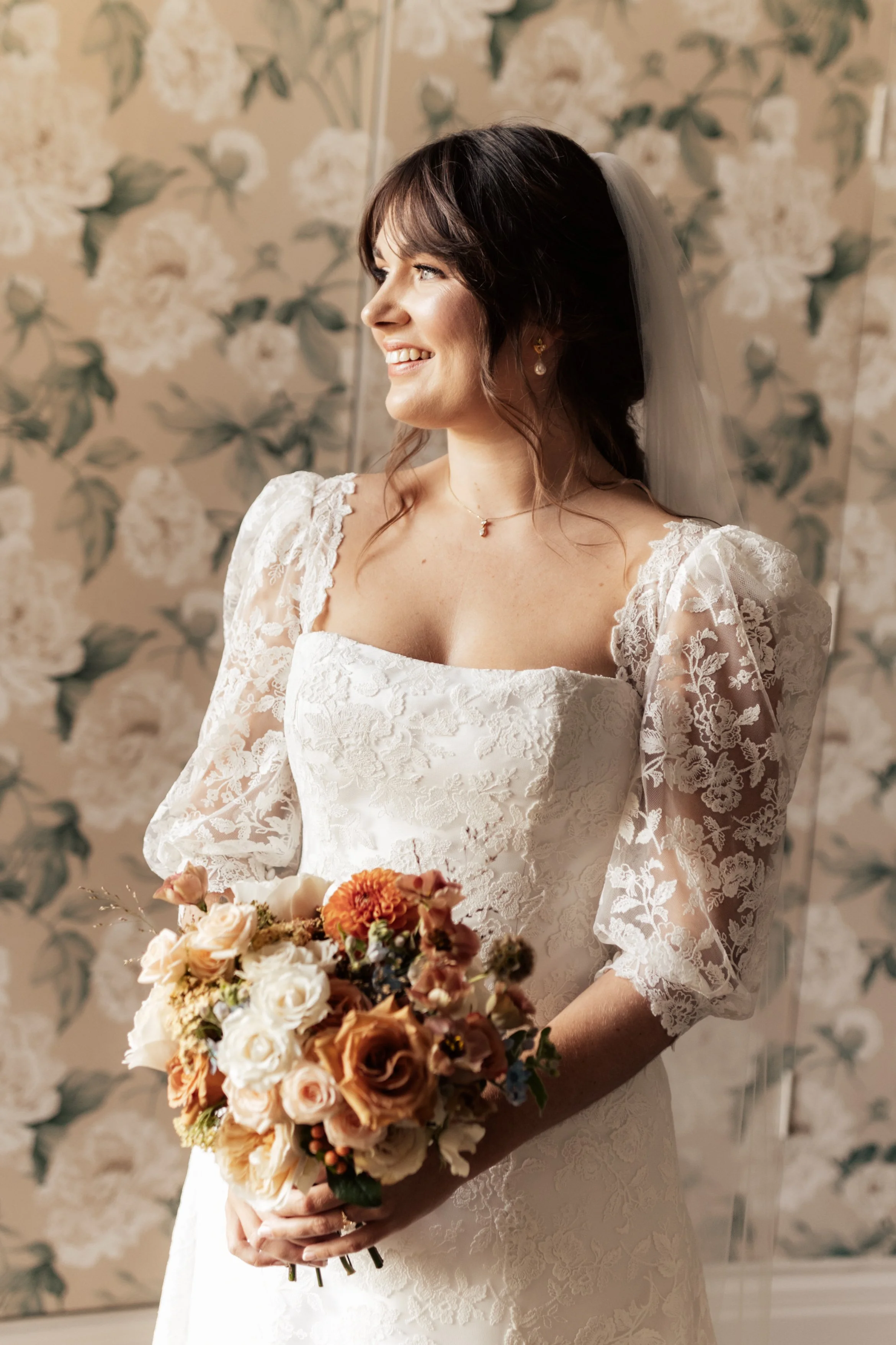 A bride with dark hair in a lace wedding dress, holding a bouquet of flowers, smiling, in front of a floral wallpaper background.