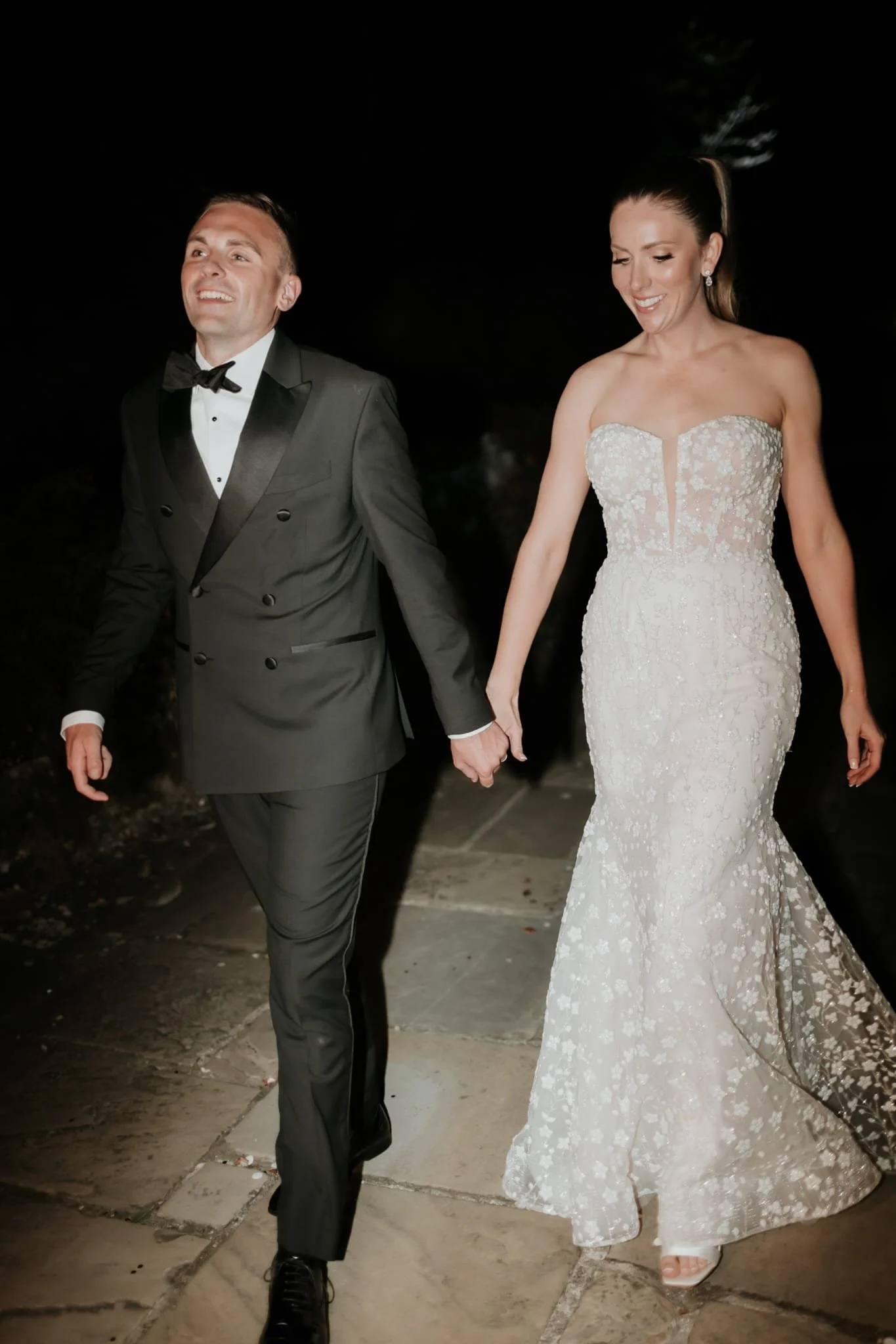 A man and woman in wedding attire walking hand-in-hand at night, smiling, with the woman in a strapless wedding dress and the man in a tuxedo.