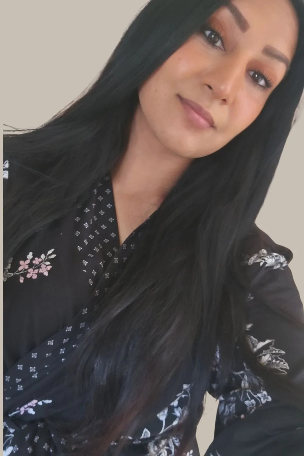 Close-up of a woman with long black hair, wearing black clothing with floral and geometric patterns, smiling softly.
