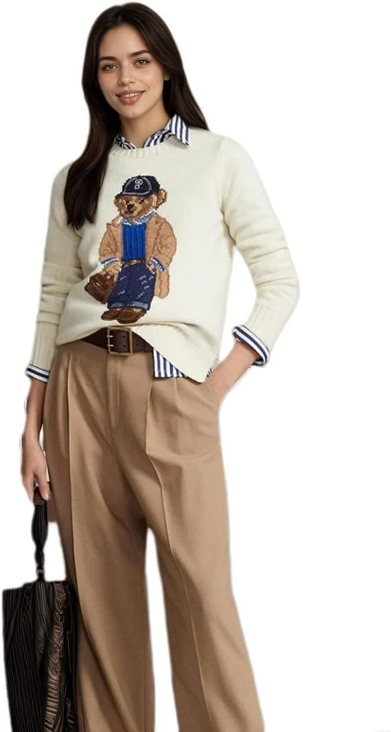 A young woman with dark hair wearing a cream sweater with a bear wearing a baseball cap and overalls, a striped shirt underneath, and tan wide-leg pants, holding a black bag, standing against a white background.