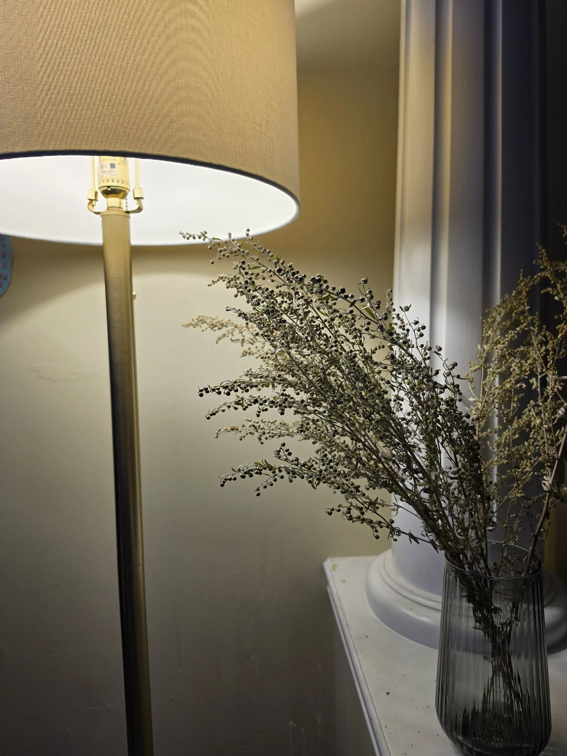 A table lamp with a beige shade, a glass vase with dried flowers, and a small white tray on a white surface.