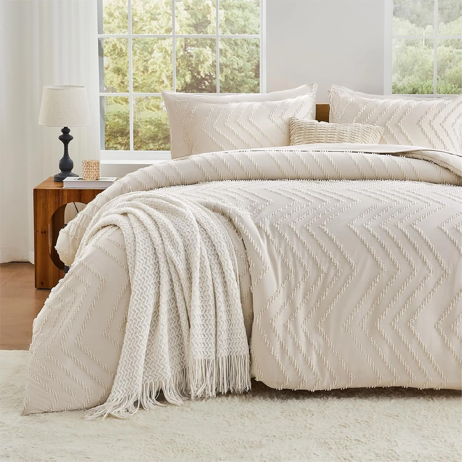 A neatly made bed with white comforter and matching pillows, a light-colored throw blanket, and a small beige decorative pillow in a bright room with large windows showing greenery outside.