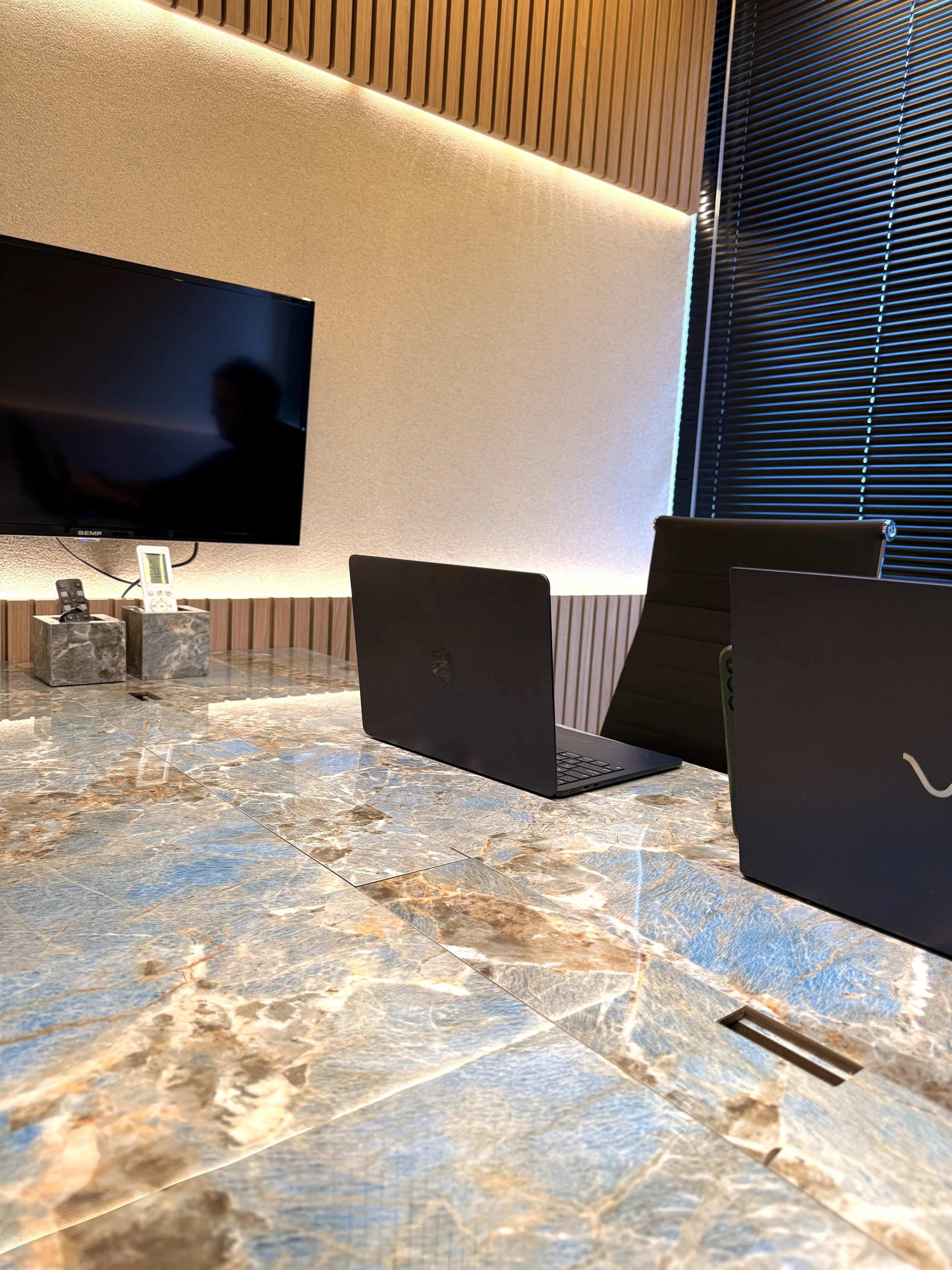 Sala de reunião com mesa de mármore, dois laptops, uma TV na parede, uma cadeira de escritório e cortinas pretas.