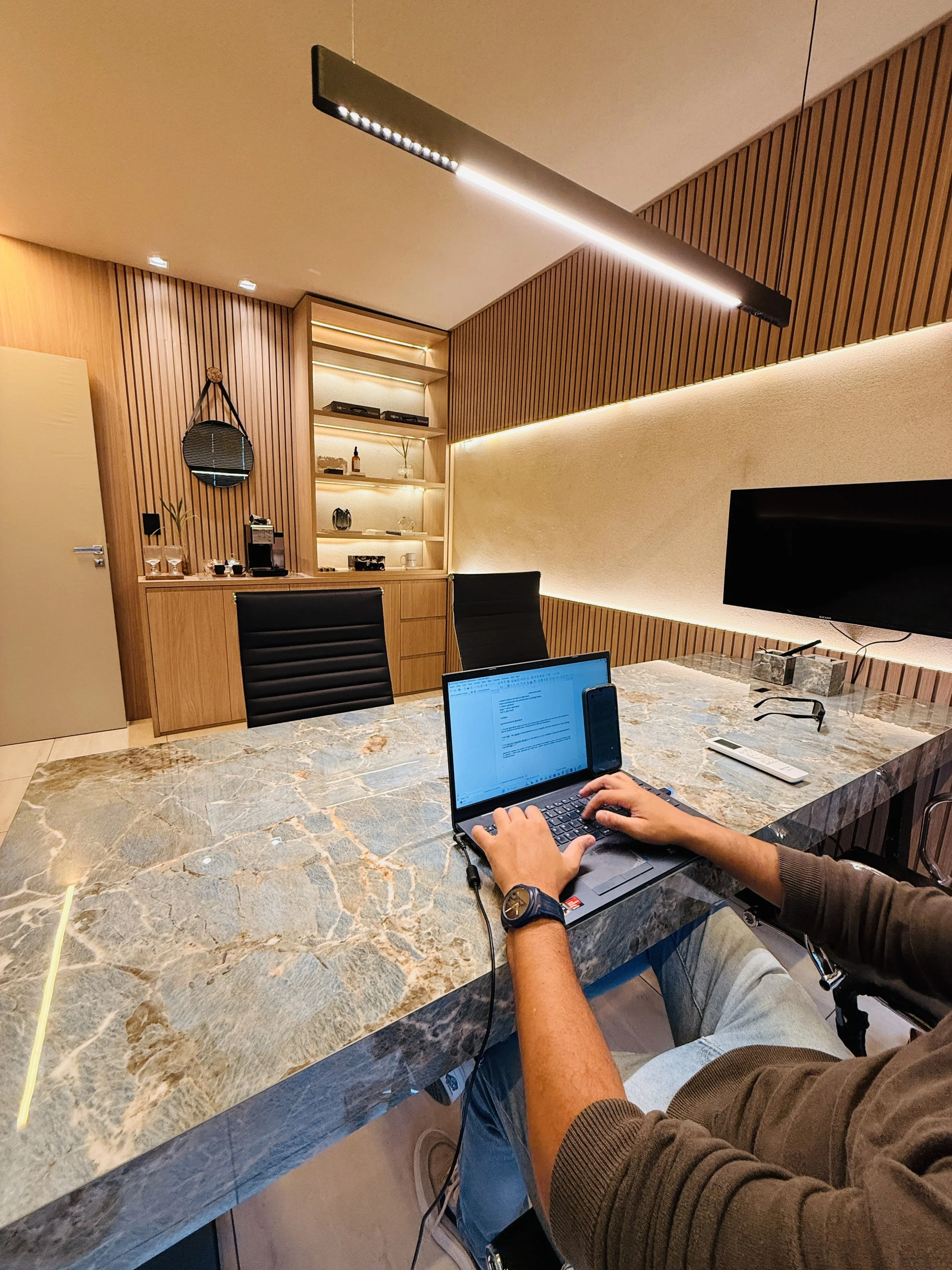 Pessoa trabalhando em uma mesa de escritório com um computador portátil em um ambiente modernizado com paredes de madeira e iluminação indireta.