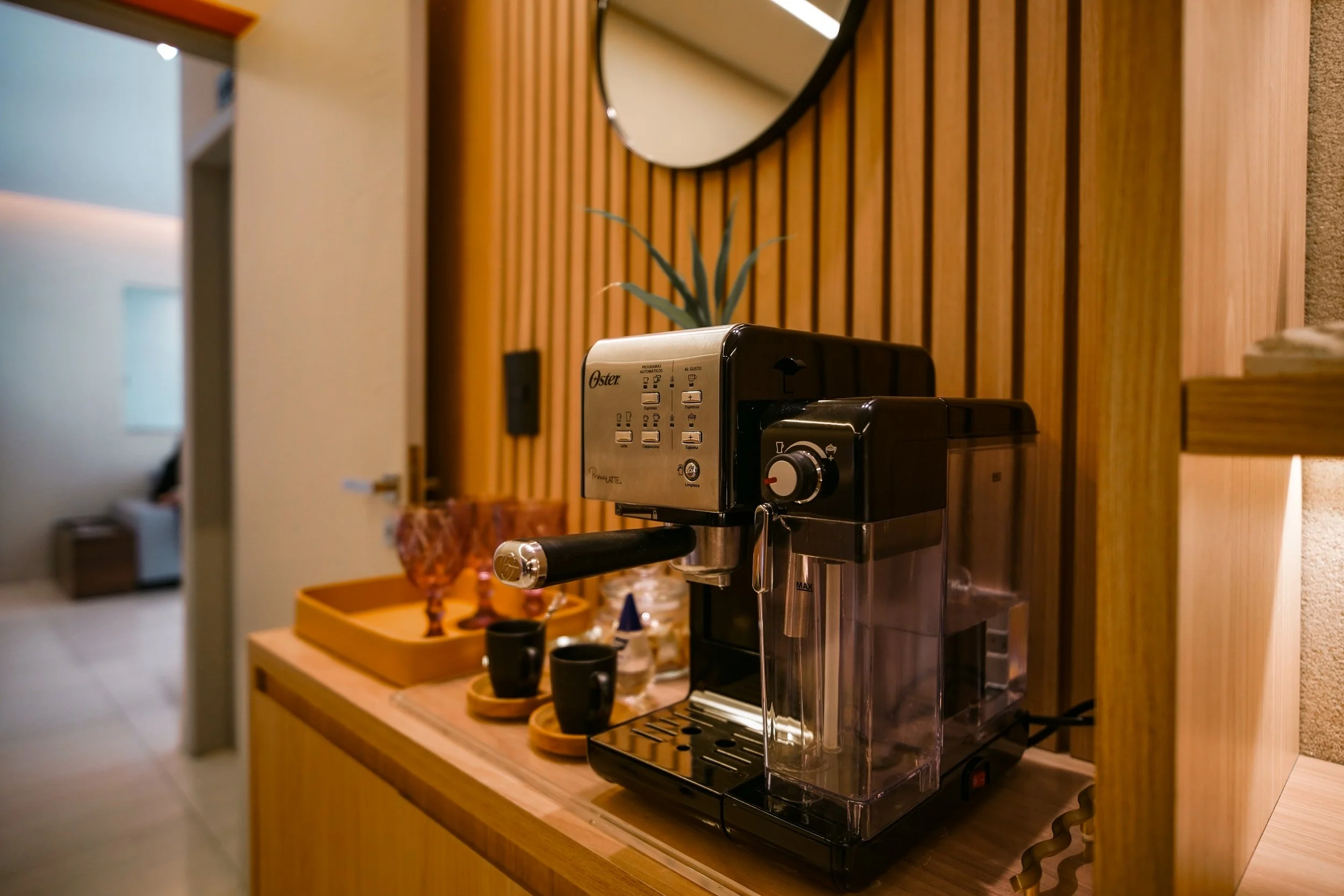 Cafeteira de café expresso em uma bancada de madeira, com duas xícaras pequenas ao lado, atrás uma bandeja com itens decorativos e uma planta.