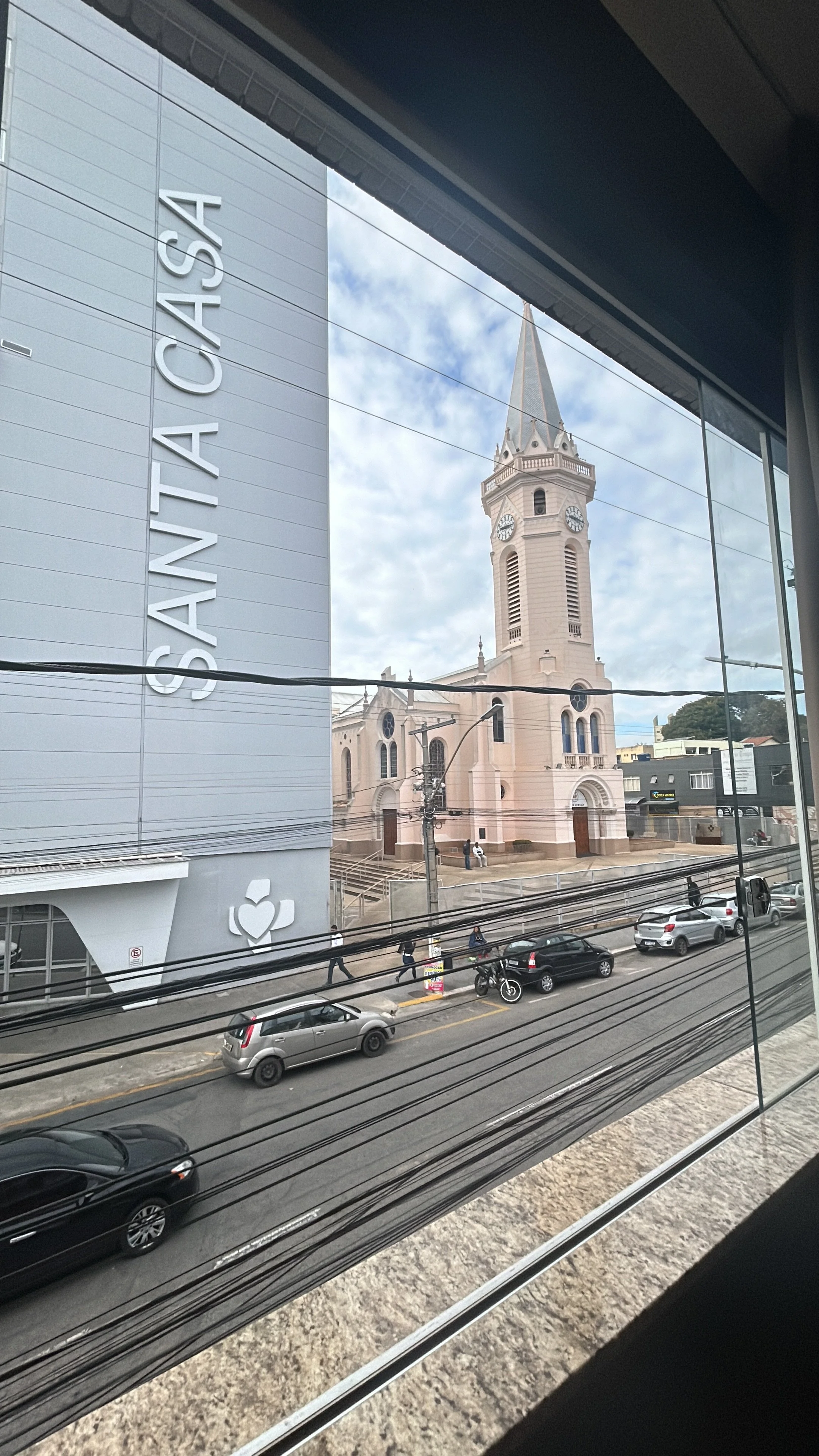 Vista da rua com carros e pessoas andando na calçada, ao fundo uma igreja com torre e relógio, ao lado um prédio moderno com a palavra 'SANTAS' na fachada.