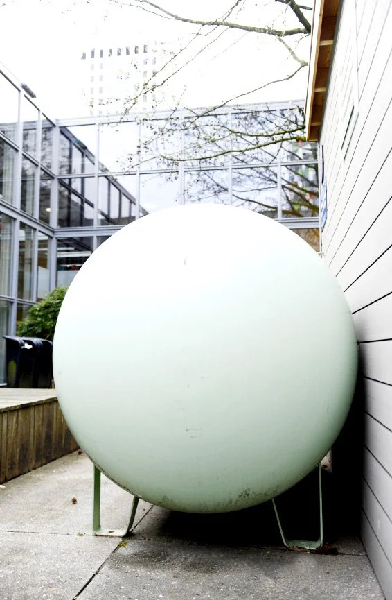 A large, round, white object on a metal stand, situated outdoors beside a house with a white siding wall, with trees and a glass building in the background.