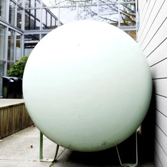 Large round white object positioned outdoors next to a white wooden wall with metal supports underneath, with a greenhouse structure and trees in the background.