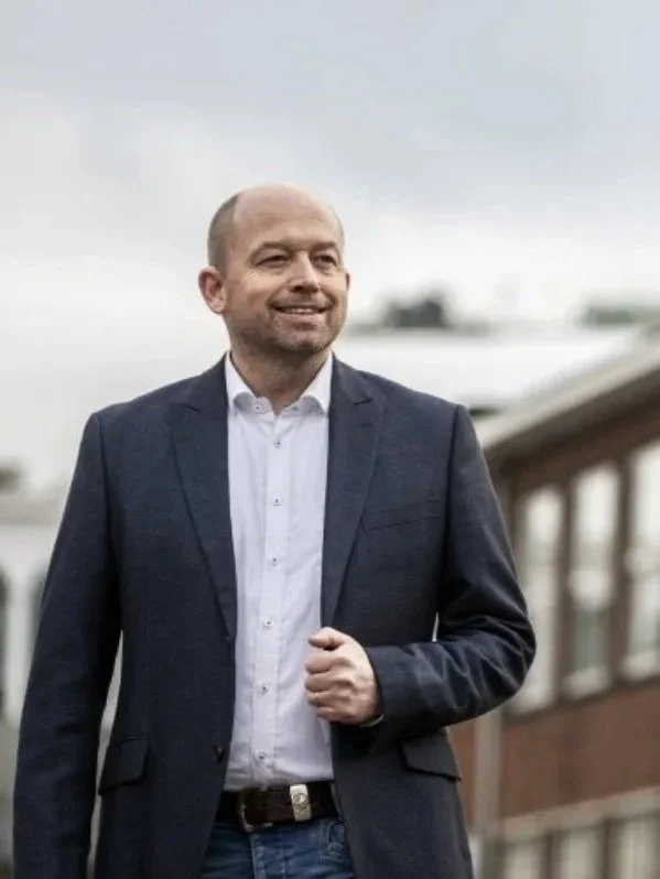 Smiling man in a suit walking outdoors, with a blurred background and overcast sky.