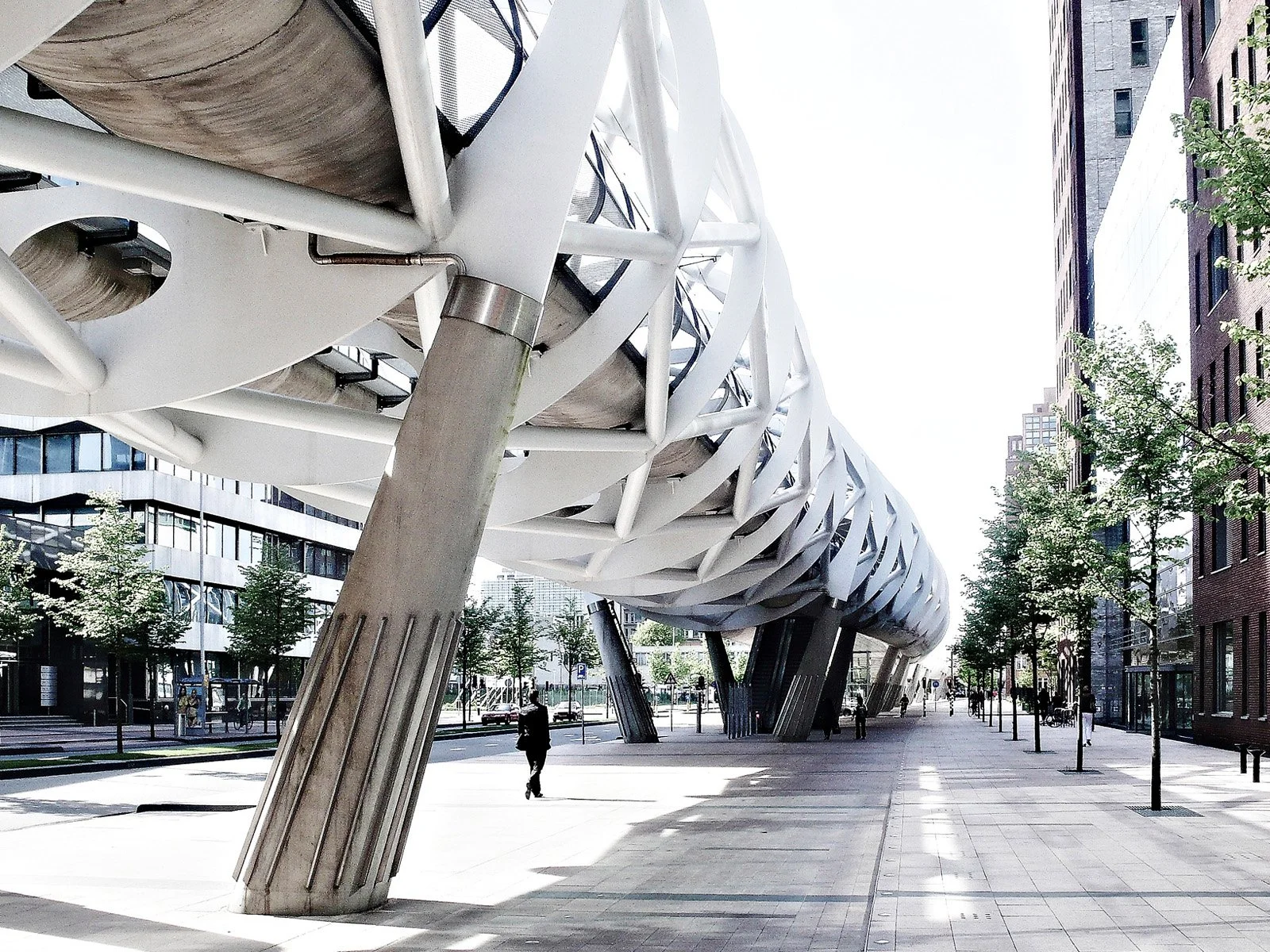 Modern architectural structure with white and black lattice design supported by angled concrete pillars, in an urban setting with tall buildings, trees, and pedestrians.