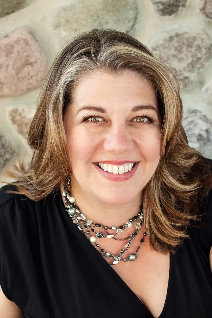 A woman with shoulder-length light brown hair smiling, wearing a black top and layered pearl and beaded necklaces, with a stone wall background.