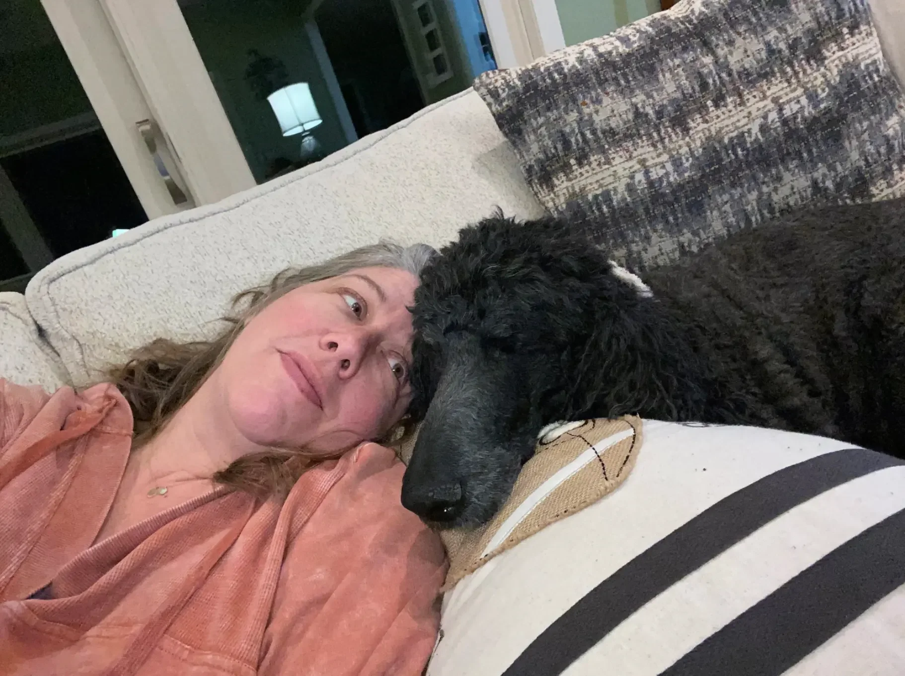 A woman with shoulder-length gray hair lying on a couch, resting her head against a cushion, with a black and white dog lying next to her, both appearing relaxed and close.
