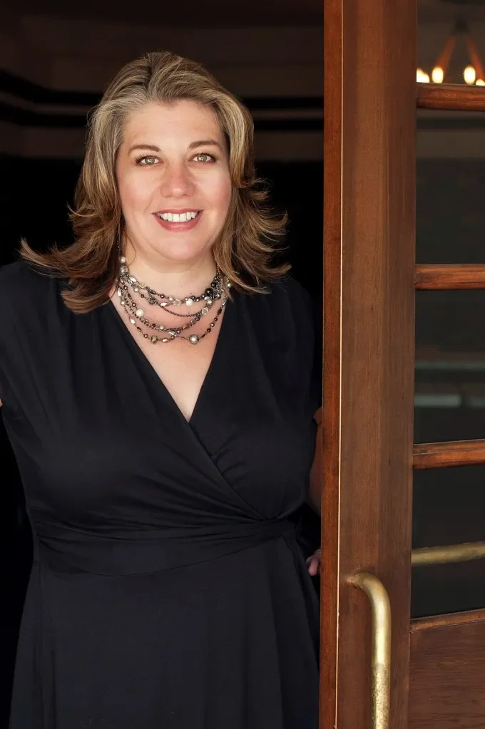 A woman with blonde hair, wearing a black dress and layered pearl necklaces, smiling while standing next to a wooden door with a brass handle.