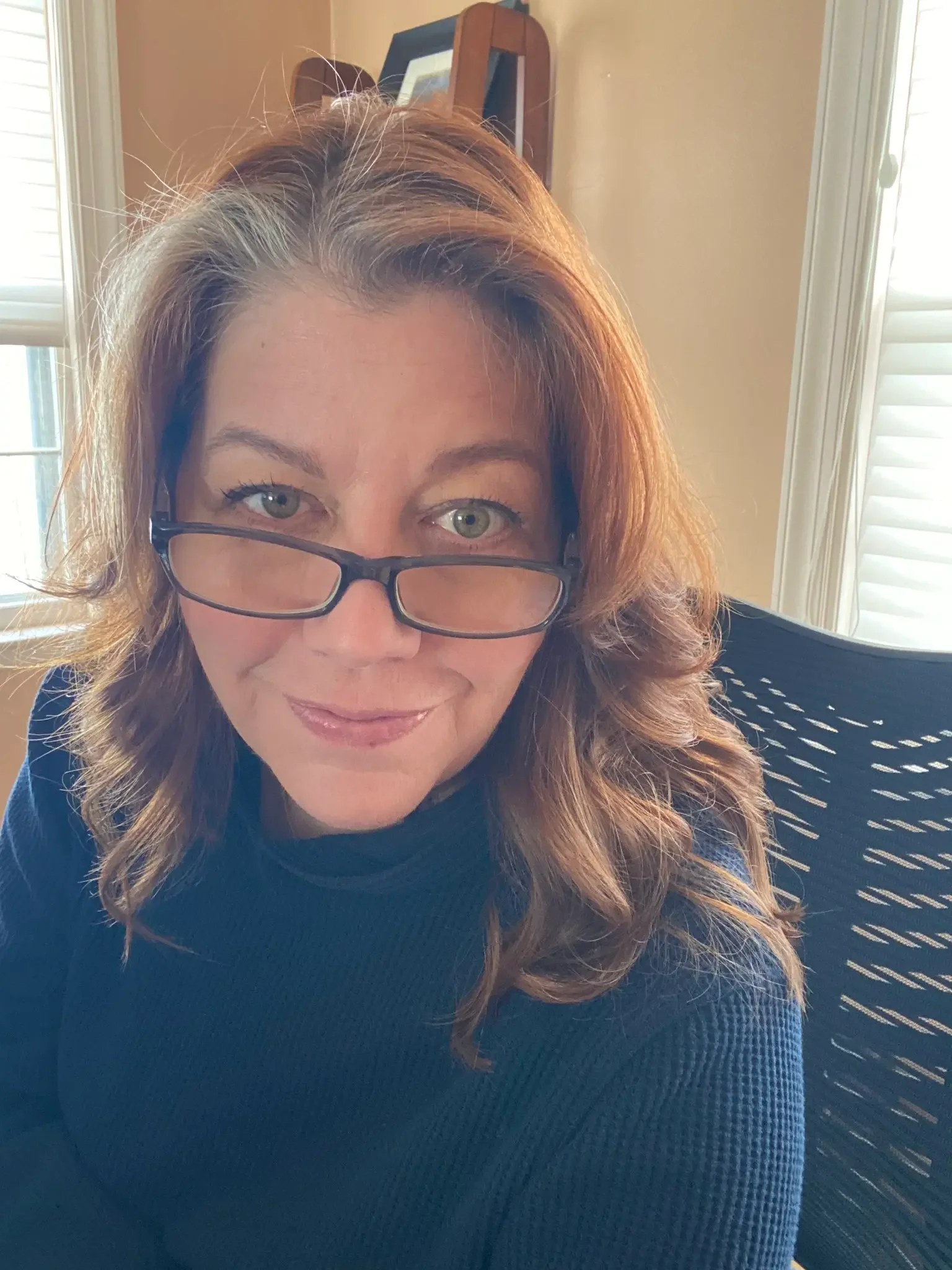 A woman with light brown, wavy hair and light blue eyes, wearing glasses perched on her nose, sitting indoors near a window with white blinds, with a framed picture on a wooden shelf behind her, smiling slightly at the camera.