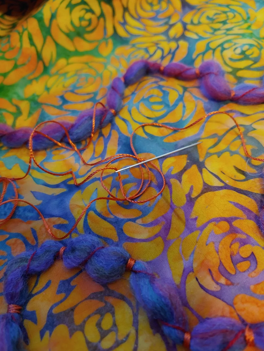 Colorful fabric with yellow, green, blue, and orange floral pattern, decorated with a purple felted necklace with round beads, along with orange and red threads and a sewing pin.