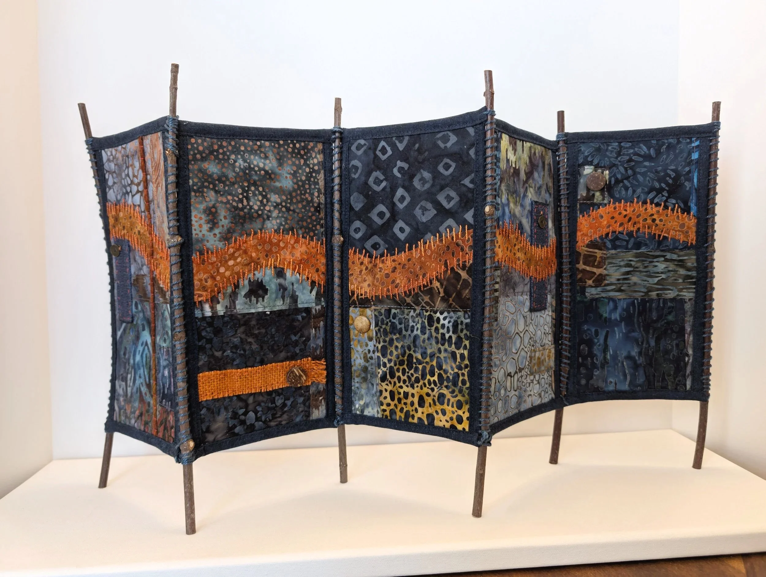 A fabric art piece with multicolored patches and embroidered orange wave designs, supported by a frame with wooden sticks, displayed on a white pedestal.