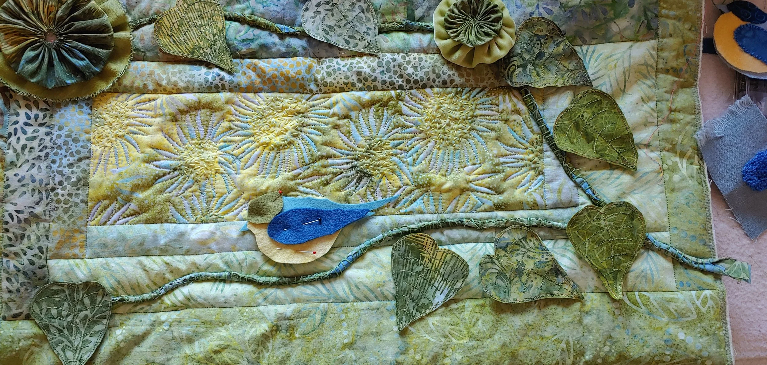 Quilted textile art featuring a branch with green leaves and a small blue bird perched on a branch against a yellow background with quilted sunburst patterns.