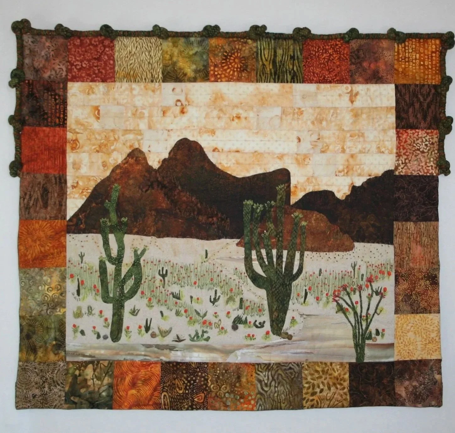 A desert-themed quilt with a landscape scene of mountains, cacti, and desert plants framed by a patchwork border of multicolored fabric squares.