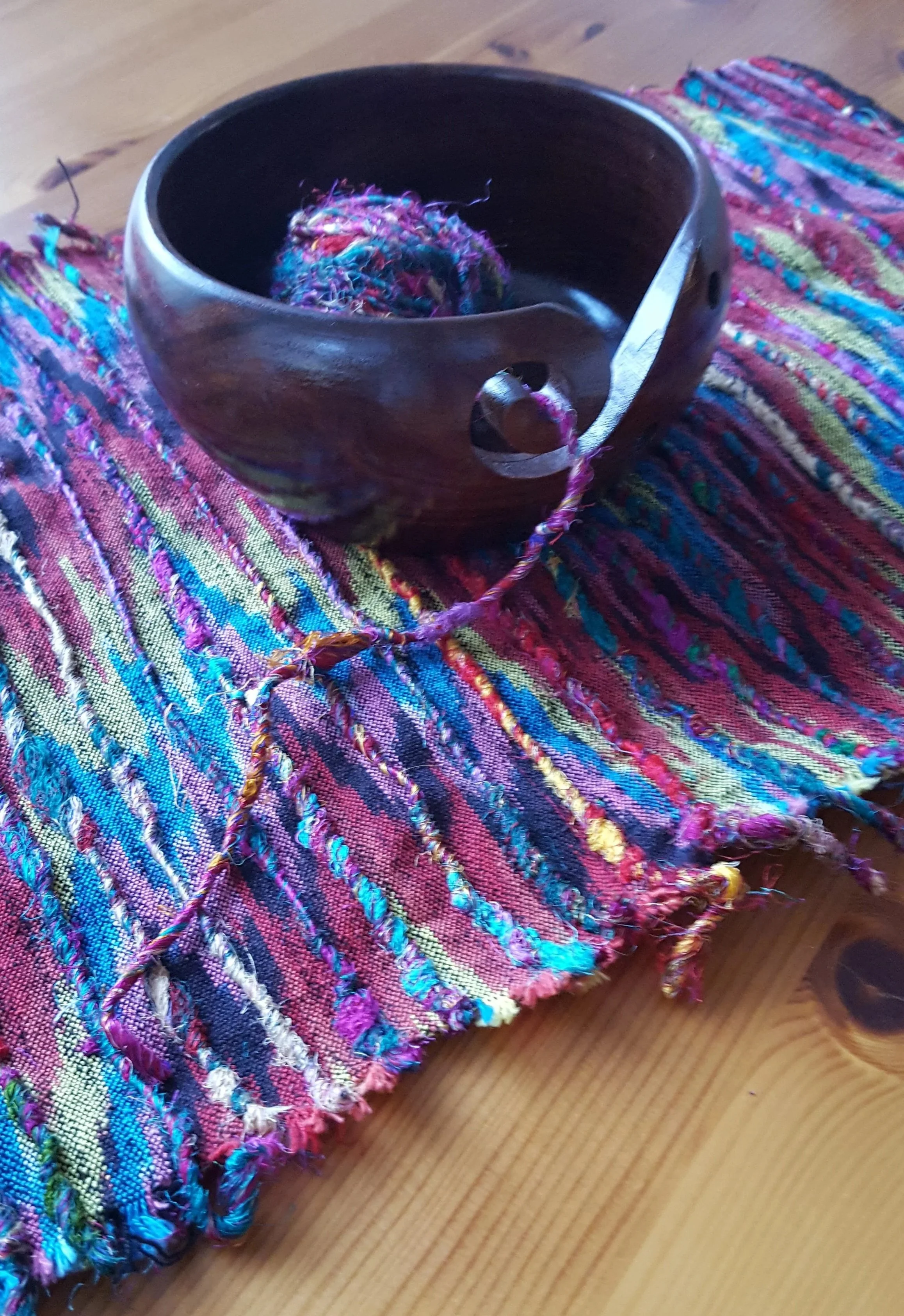 A wooden spinning top with a ball of multicolored yarn on top, positioned on a colorful woven textile on a wooden surface.