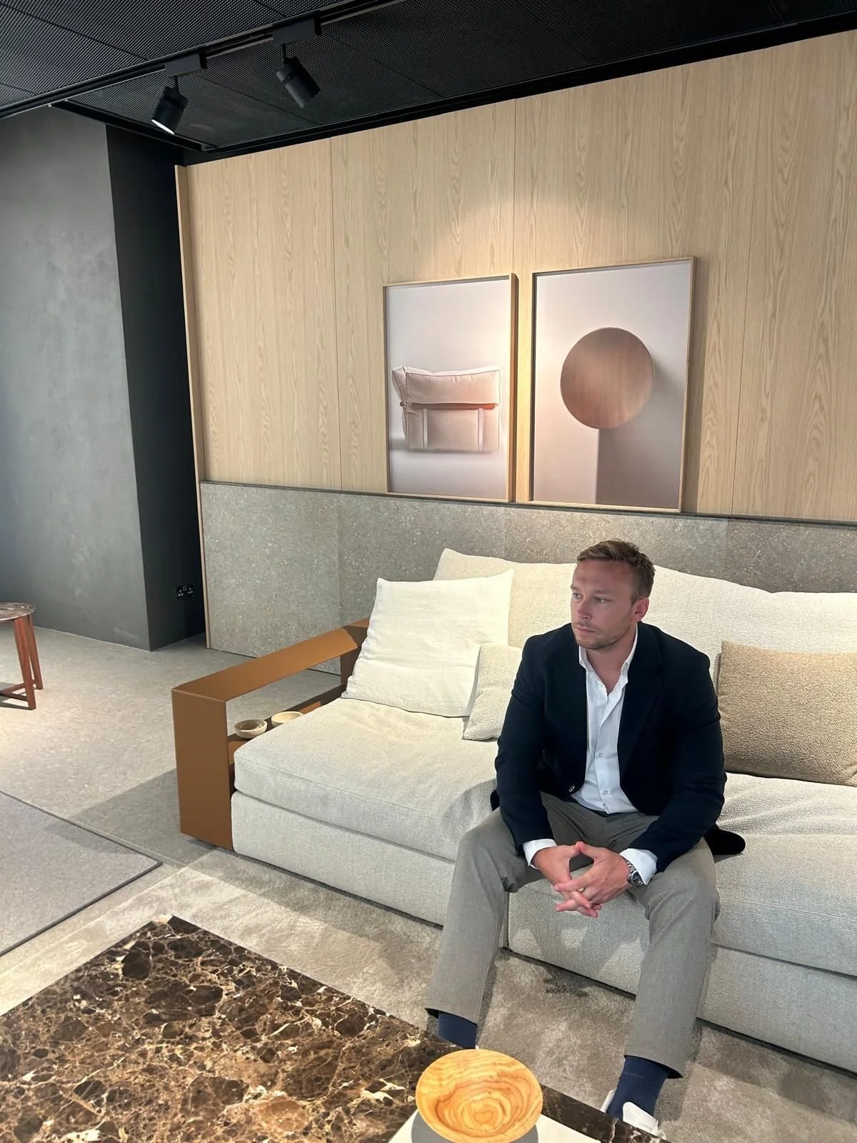 A man in business attire sitting on a light-colored sofa in a modern, minimalistic interior with wooden wall panels, abstract artwork, a marble table, and neutral decor.